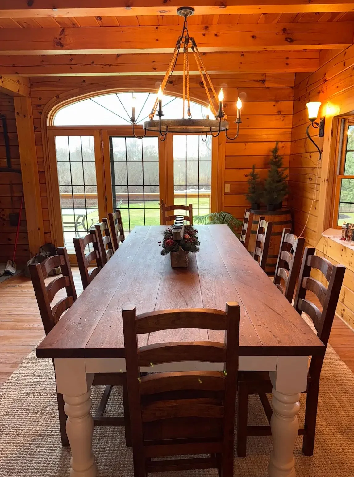 A cozy wooden dining room with a large table, ten chairs, a chandelier, and a view of a grassy yard through large windows and a sliding glass door.