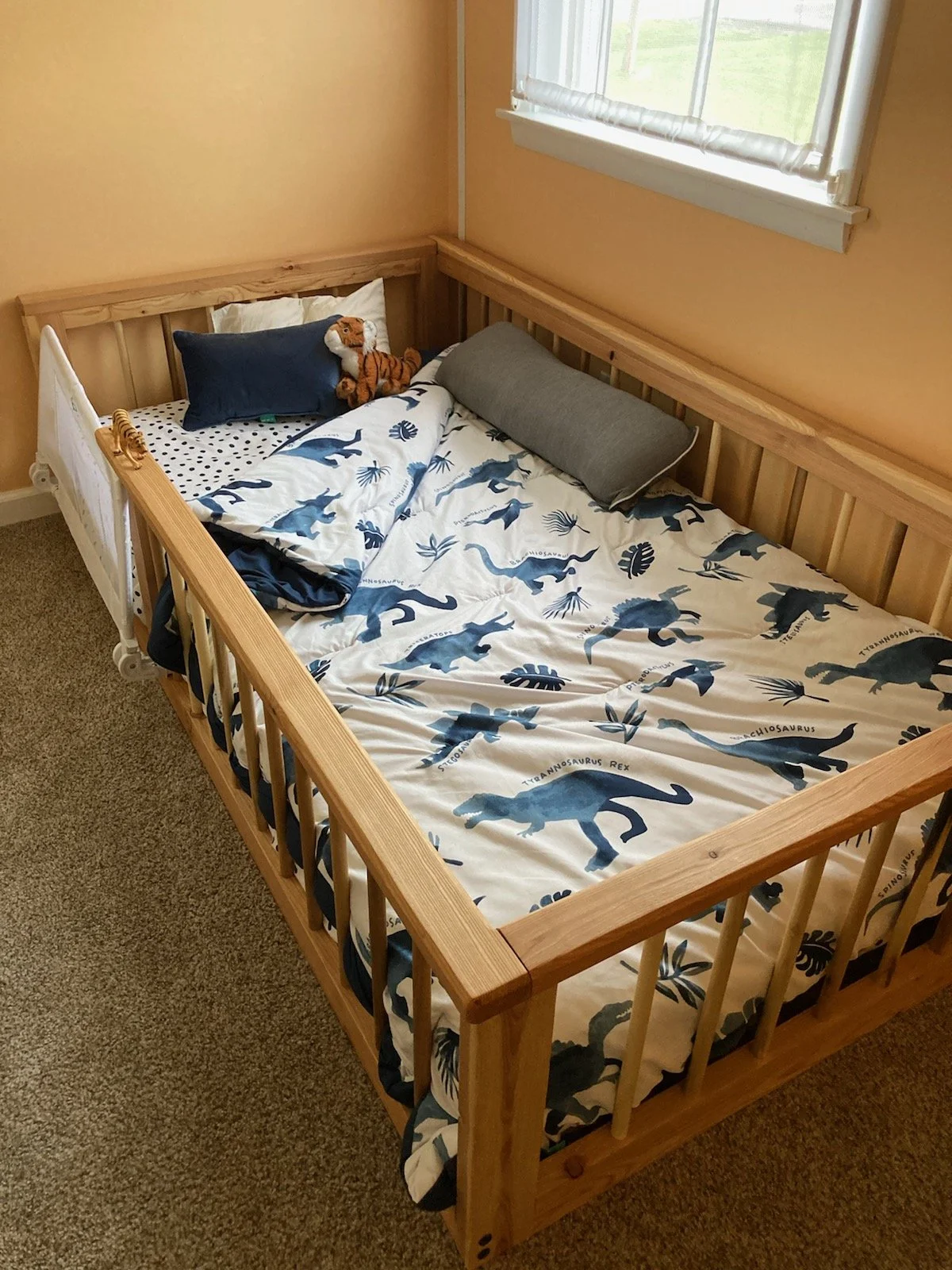 A child's bed with dinosaur-themed bedding, a tiger stuffed animal, a blue pillow, and a long gray bolster pillow, situated near a window with blinds.
