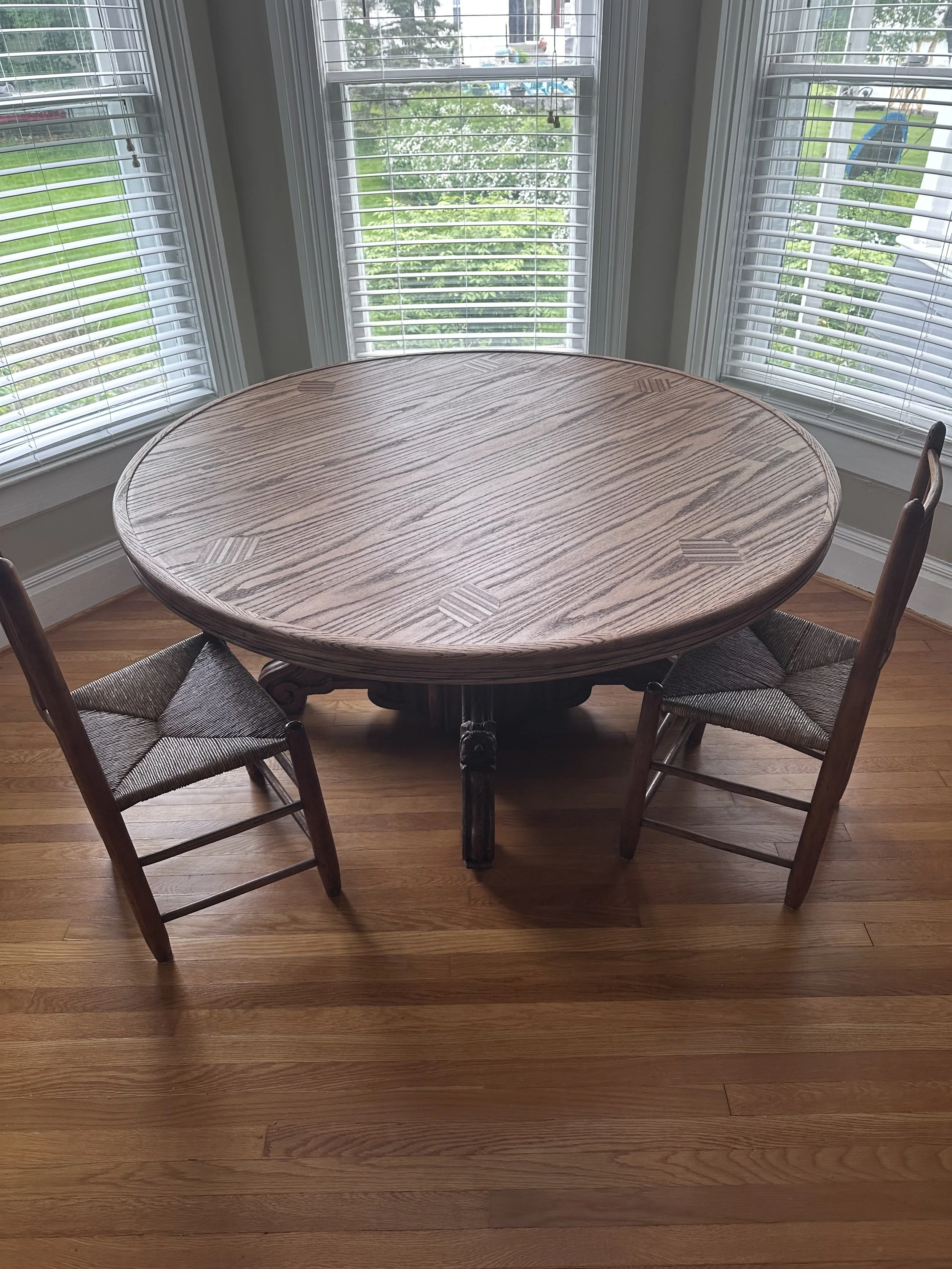 A round wooden dining table with two wooden chairs in front of a bay window with white blinds, overlooking a green lawn.