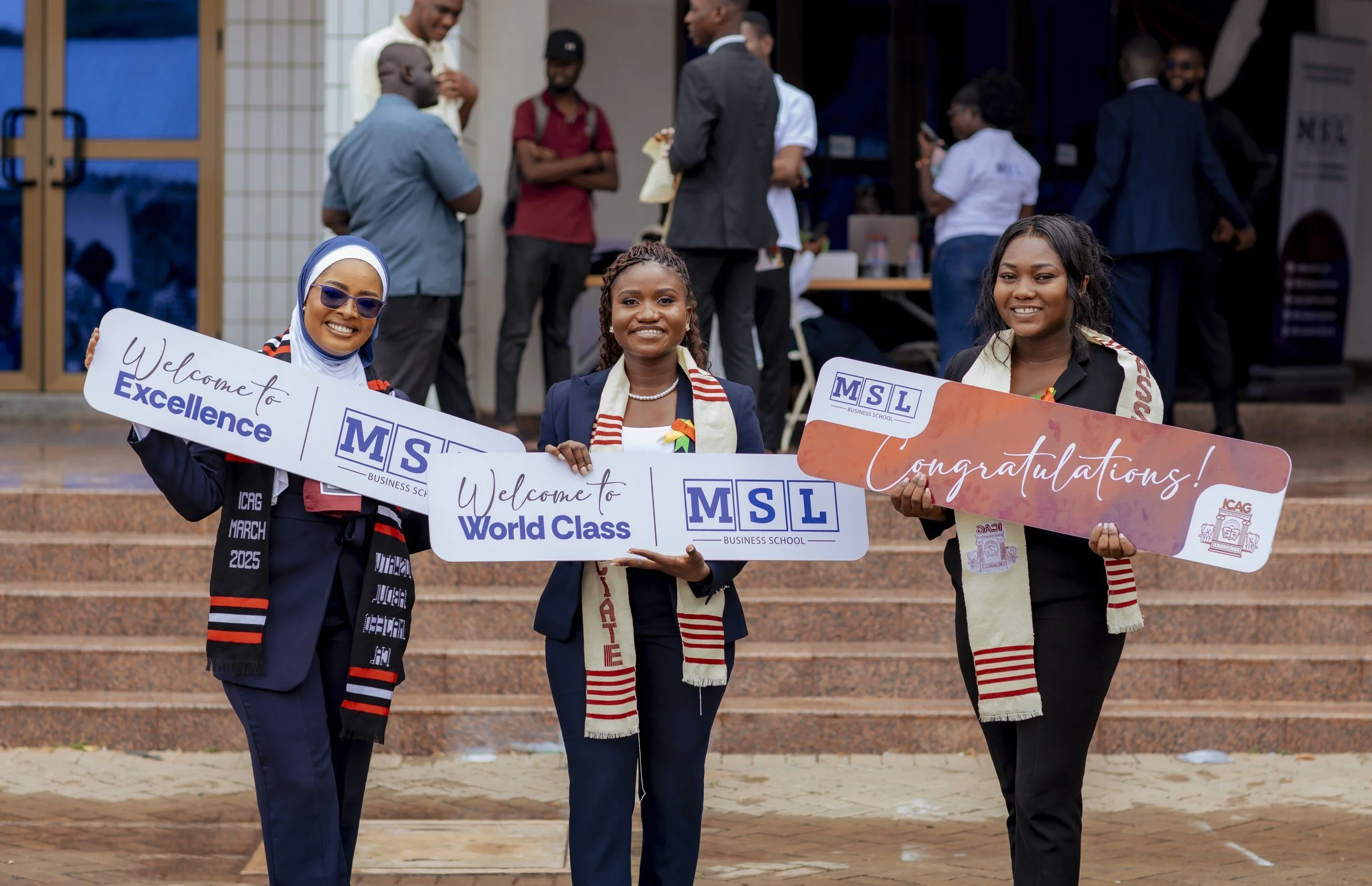 Three graduates holding MSL Welcome to Excellence, World Class and Congratulations signs on the steps, ICAG graduation, May 2025.