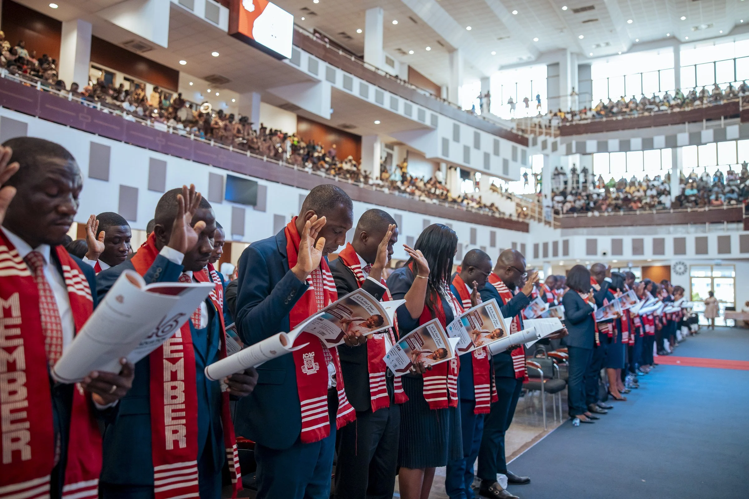 Graduates in red ICAG stoles reading ceremony sheets in a row beside the stage, ICAG graduation, February 2026.