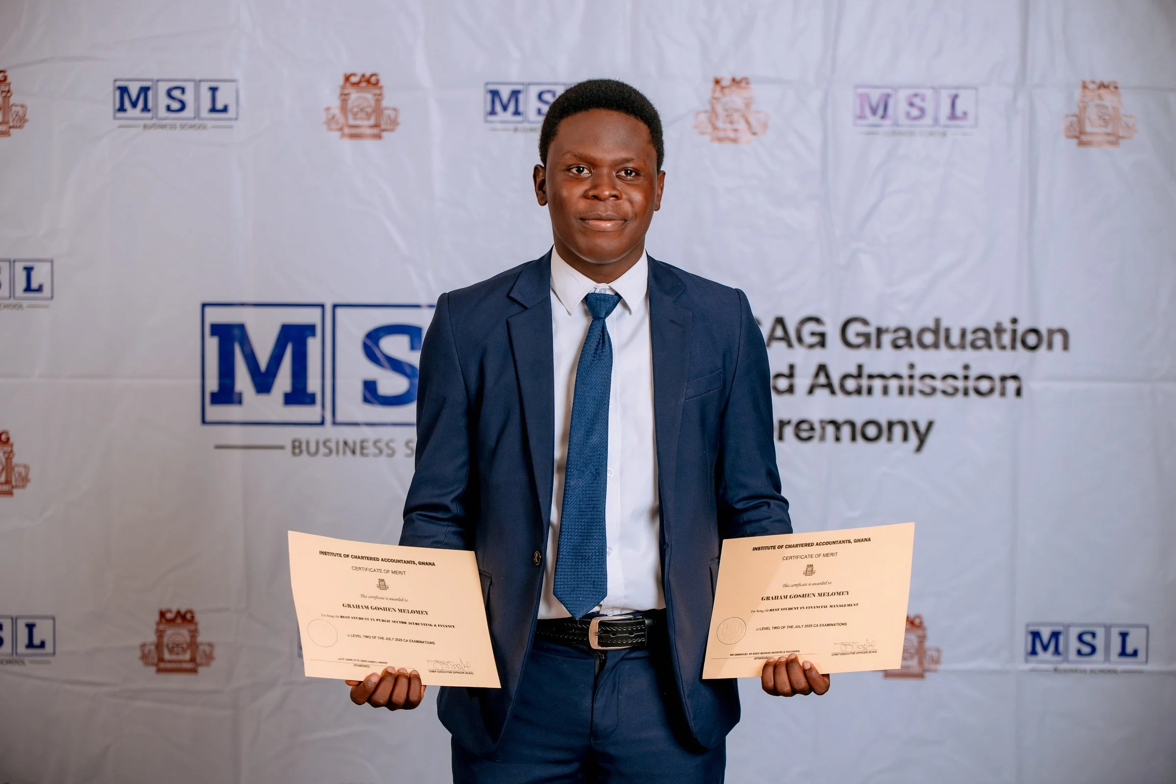 Smiling graduate in a navy suit displaying two certificates at the MSL backdrop, ICAG graduation, September 2025.