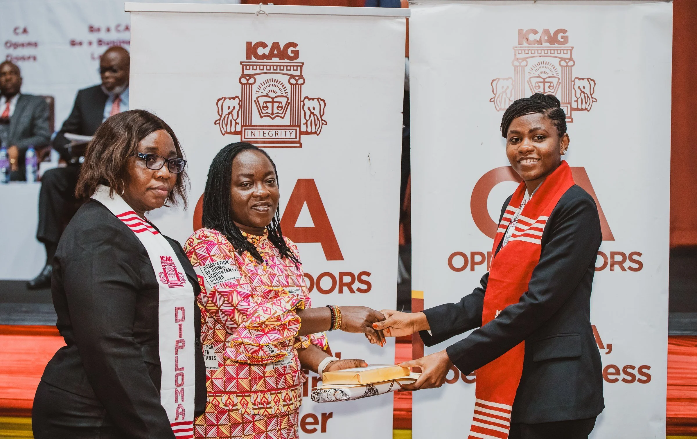 Graduate in a red ICAG stole receiving her certificate from two women on stage, ICAG graduation, September 2025.