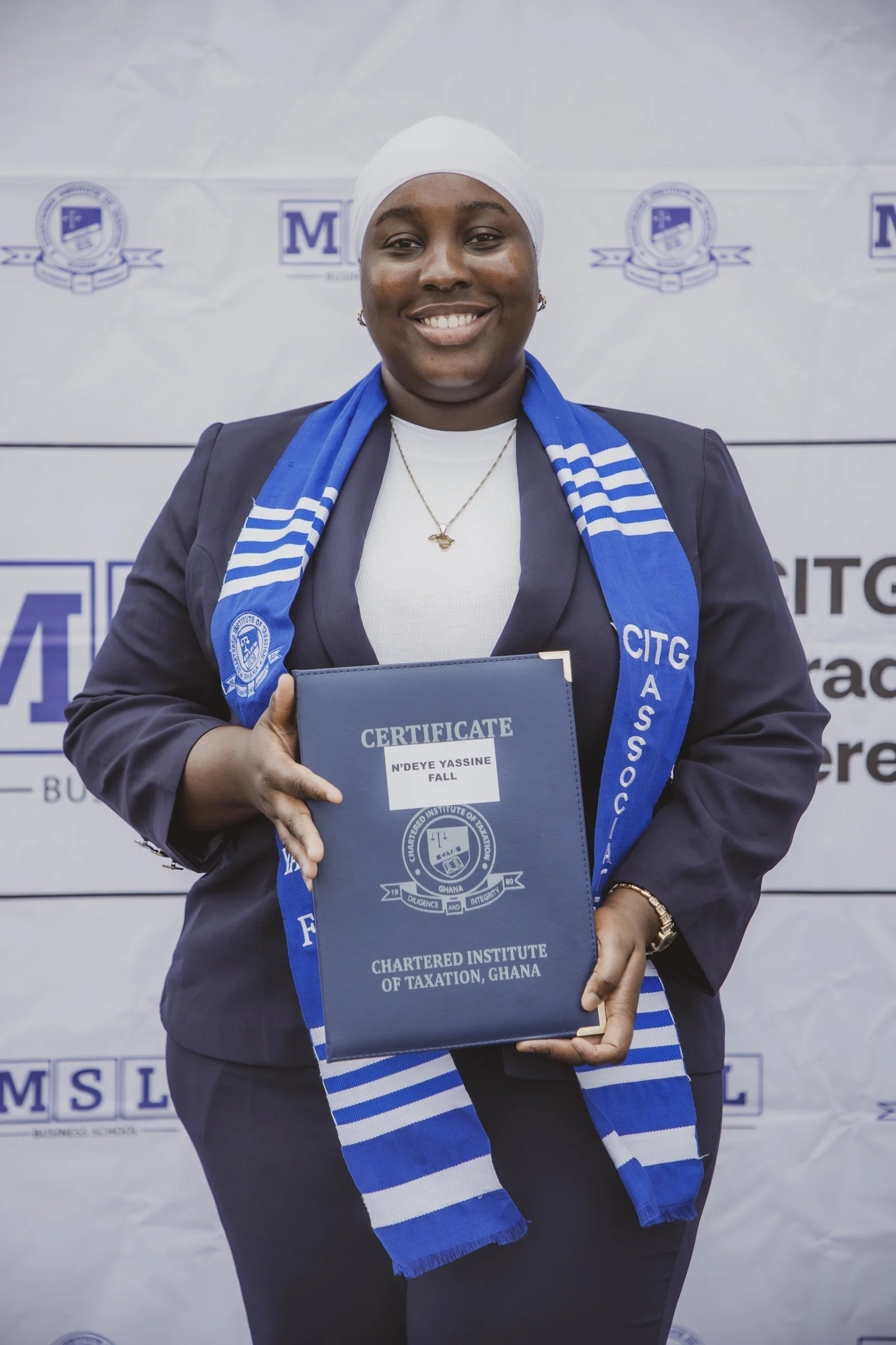 Smiling graduate in a white headscarf and blue CITG stole displaying her certificate folder, CITG graduation, May 2026.