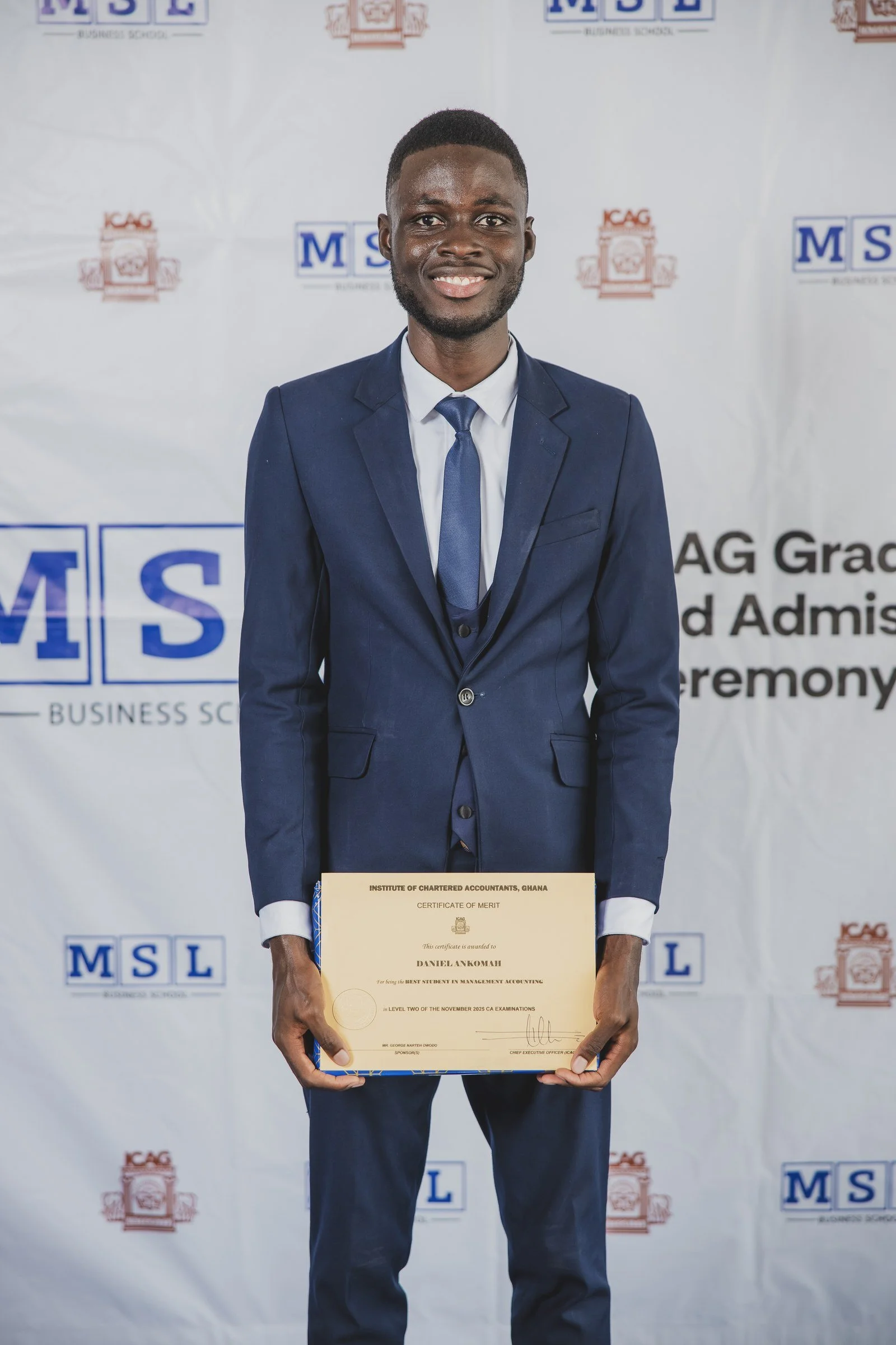 Smiling graduate wearing glasses and a navy suit holding a certificate, ICAG graduation, February 2026.
