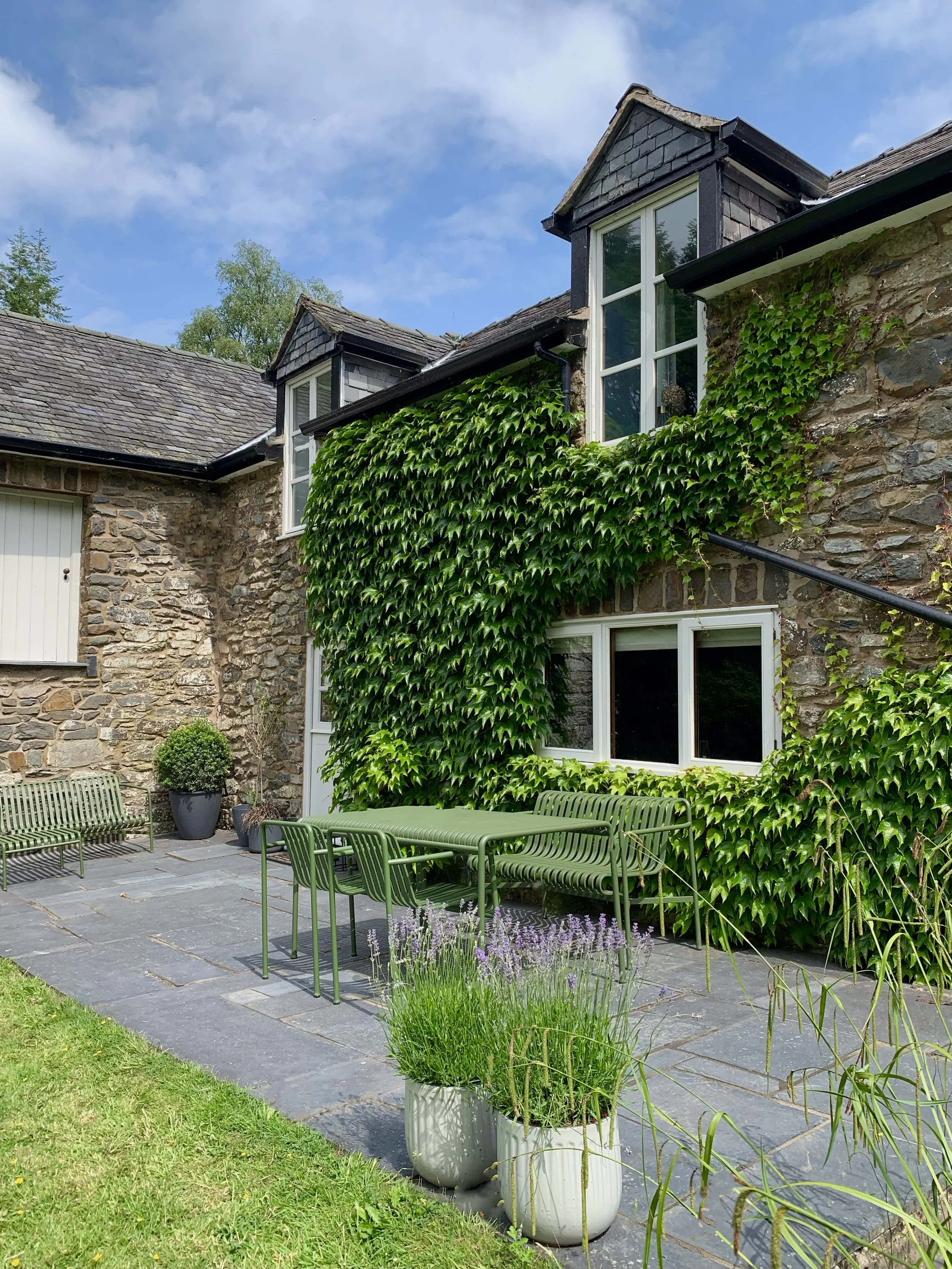 Stone house covered in ivy with a patio featuring green outdoor furniture and plants in pots.