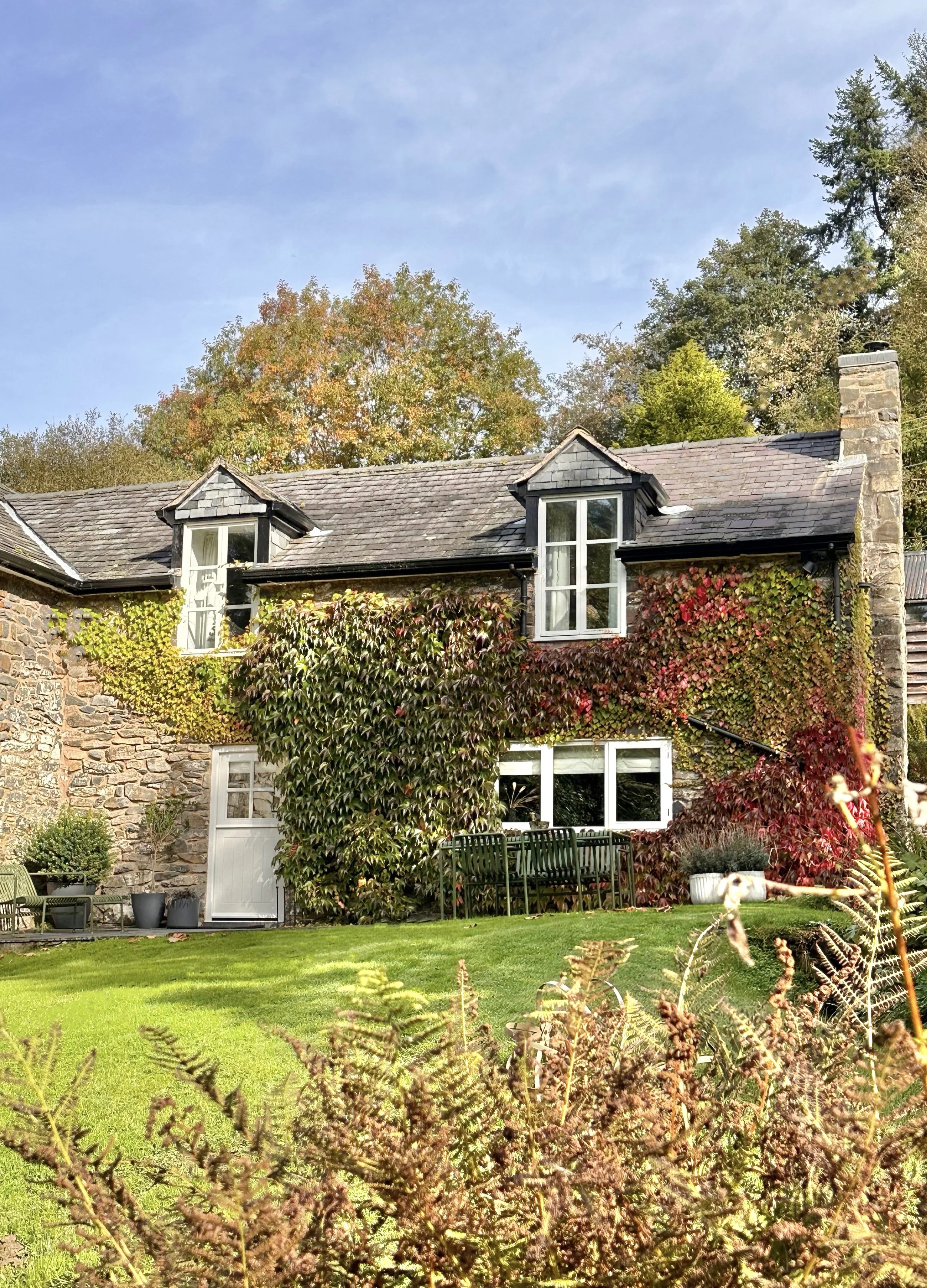Stone cottage with ivy-covered walls, two dormer windows, and a chimney, surrounded by a lush green lawn and plants, with trees in the background.