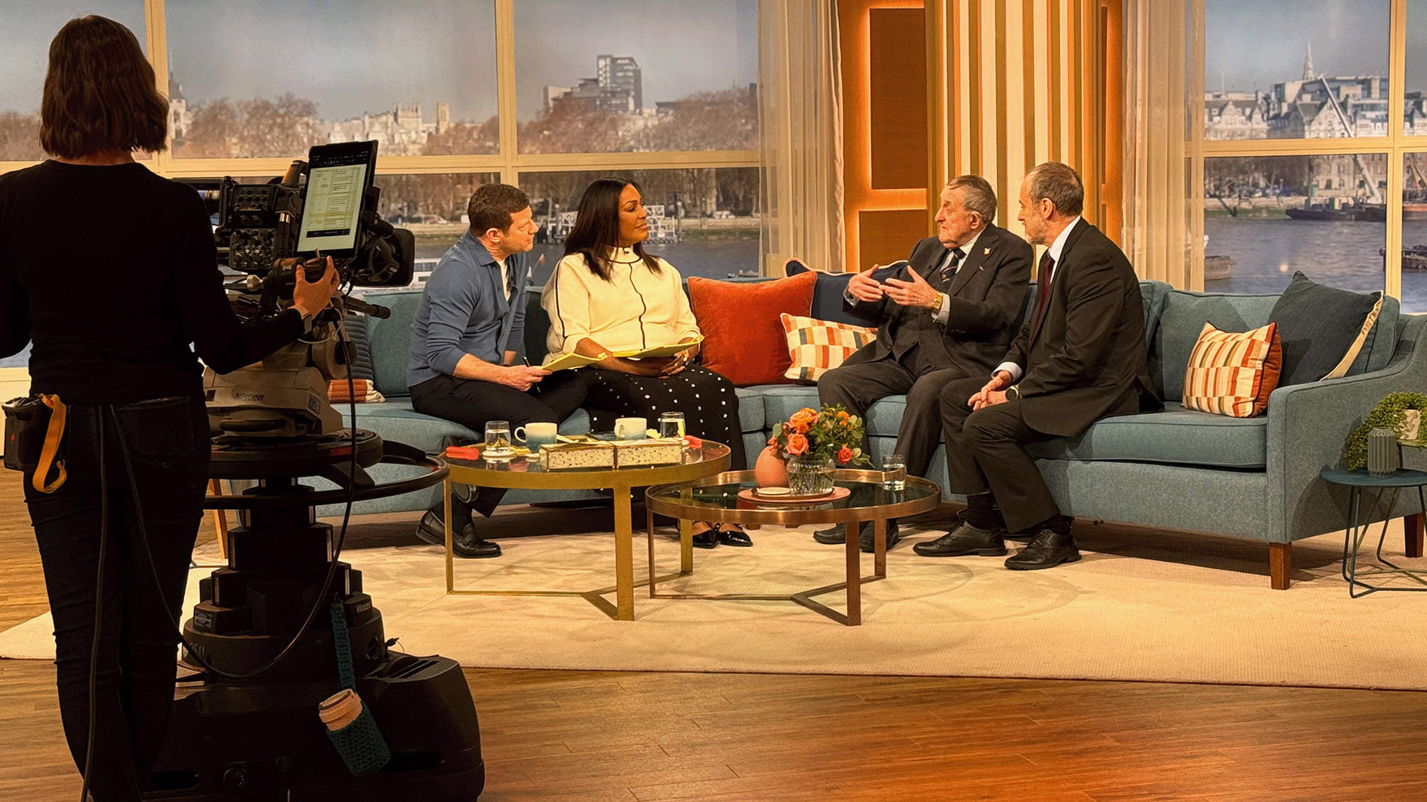 Television talk show set with five people seated on a blue sofa. A woman is holding papers, two men and a woman are engaged in conversation, and a camera operator is filming. Large windows behind them reveal a river and cityscape.