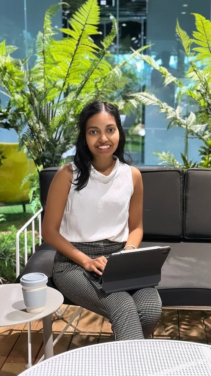 A woman sitting on a black outdoor sofa with a laptop on her lap, smiling, with green plants and glass windows in the background, and coffee cups on a small white table in front of her.