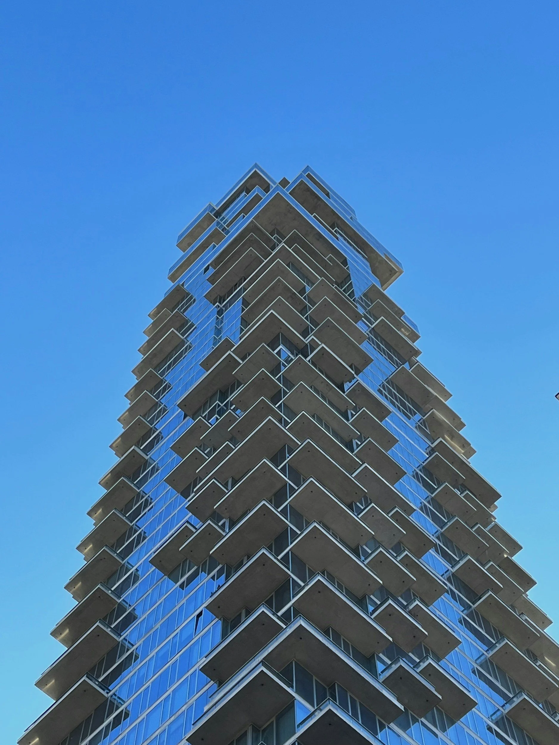 Low-angle view of a modern high-rise building with glass windows and protruding balconies against a clear blue sky.
