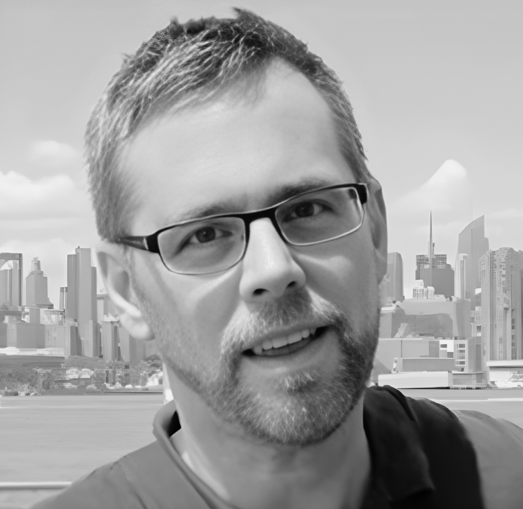 A man with glasses and a beard smiling with a city skyline in the background.