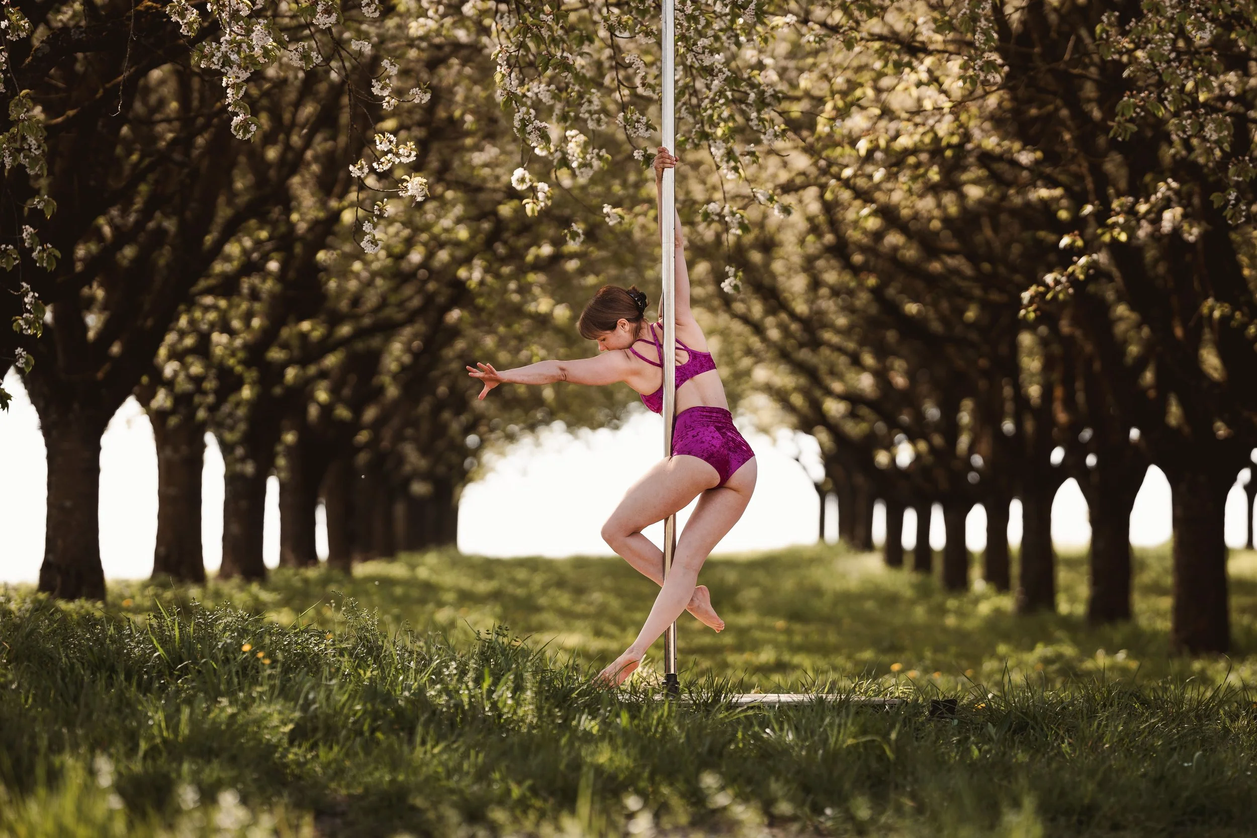 Eine Frau in pinkem Sportoutfit übt Yin-Yoga an einer Pole Dancing-Stange im Freien, umgeben von blühenden Bäumen.