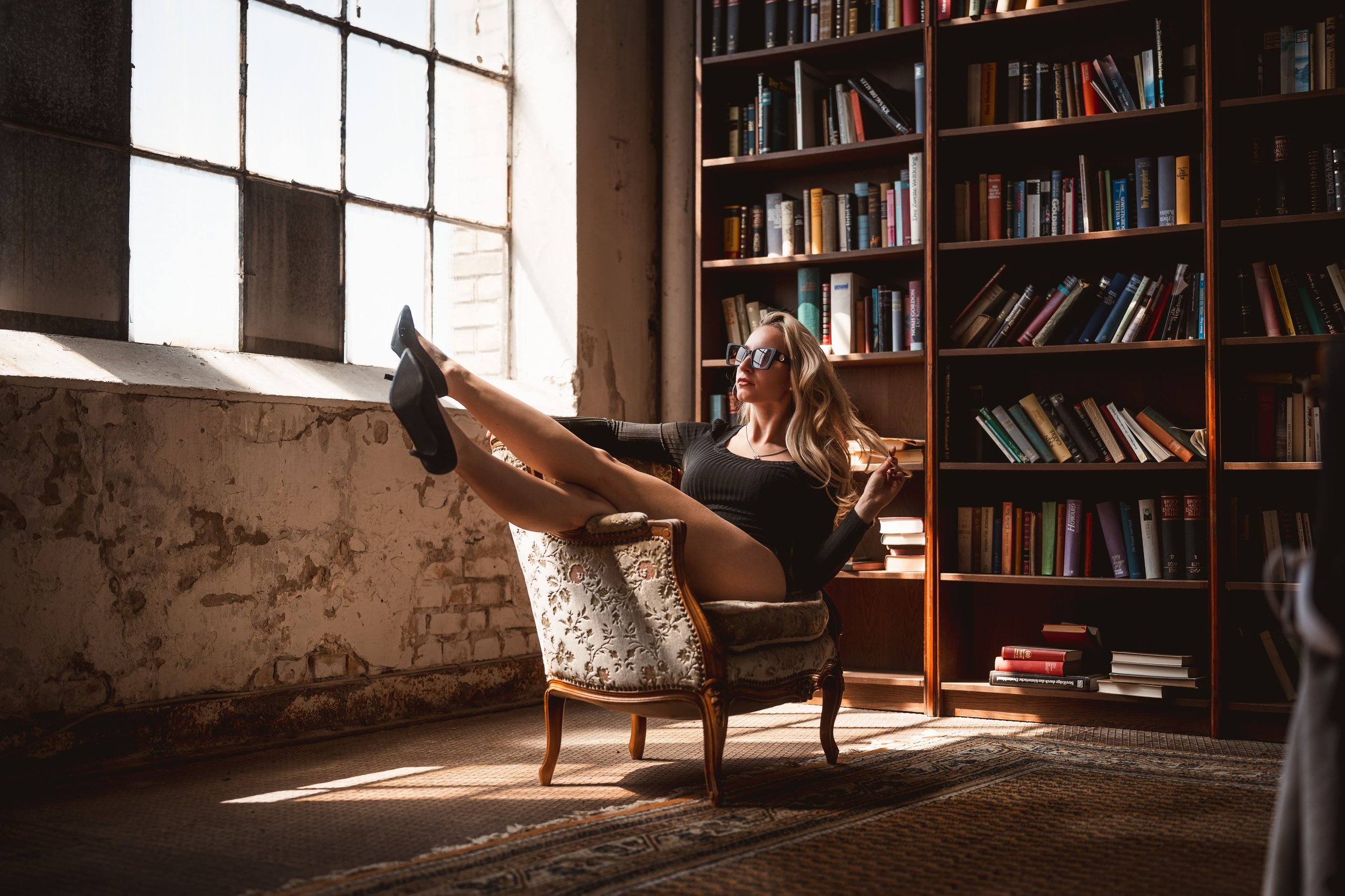 Eine junge Frau sitzt in einem stilvollen Lesezimmer an einem alten, gepolsterten Stuhl mit Holzrahmen, trägt schwarze Kleidung und Sonnenbrille, hat die Beine hochgelegt und liest ein Buch, neben ihr ein Bücherregal mit vielen bunten Büchern, das Li