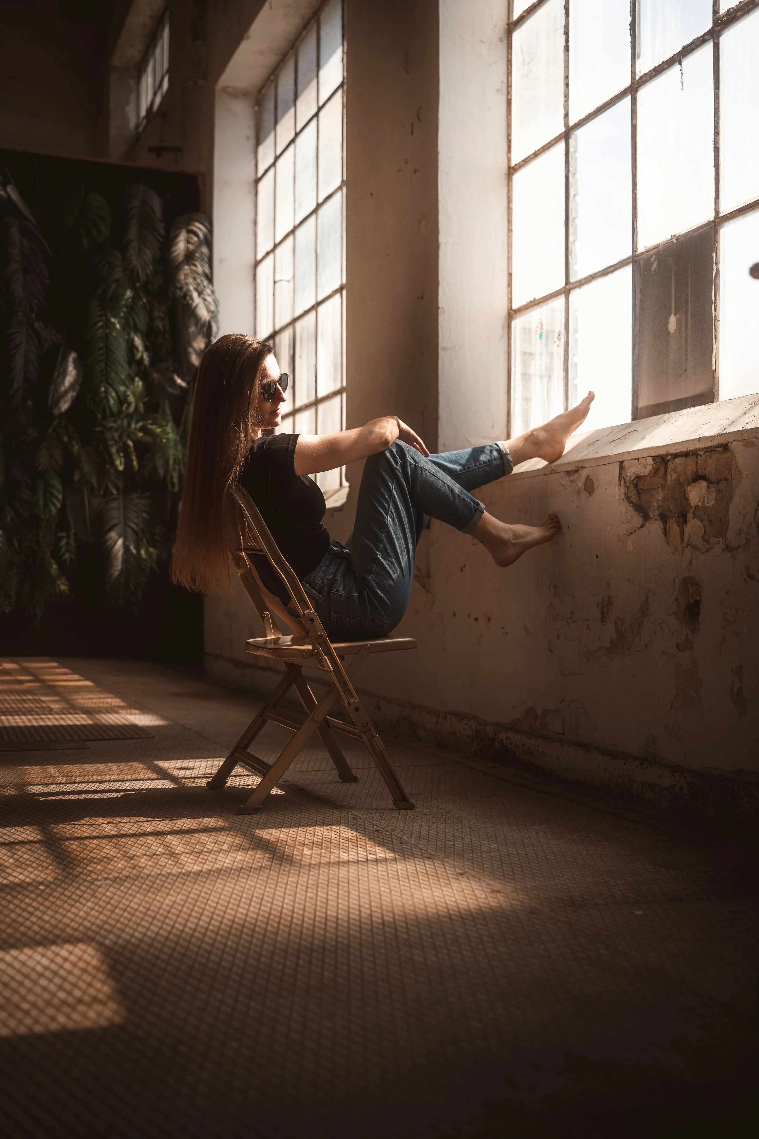 Eine junge Frau mit langen Haaren, dunklen Sonnenbrillen und casual Kleidung sitzt entspannt auf einem Klappstuhl vor einer großen Fensterwand in einem alten, industriellen Raum. Sie lehnt sich gegen die Fensterbank und blickt nach draußen.