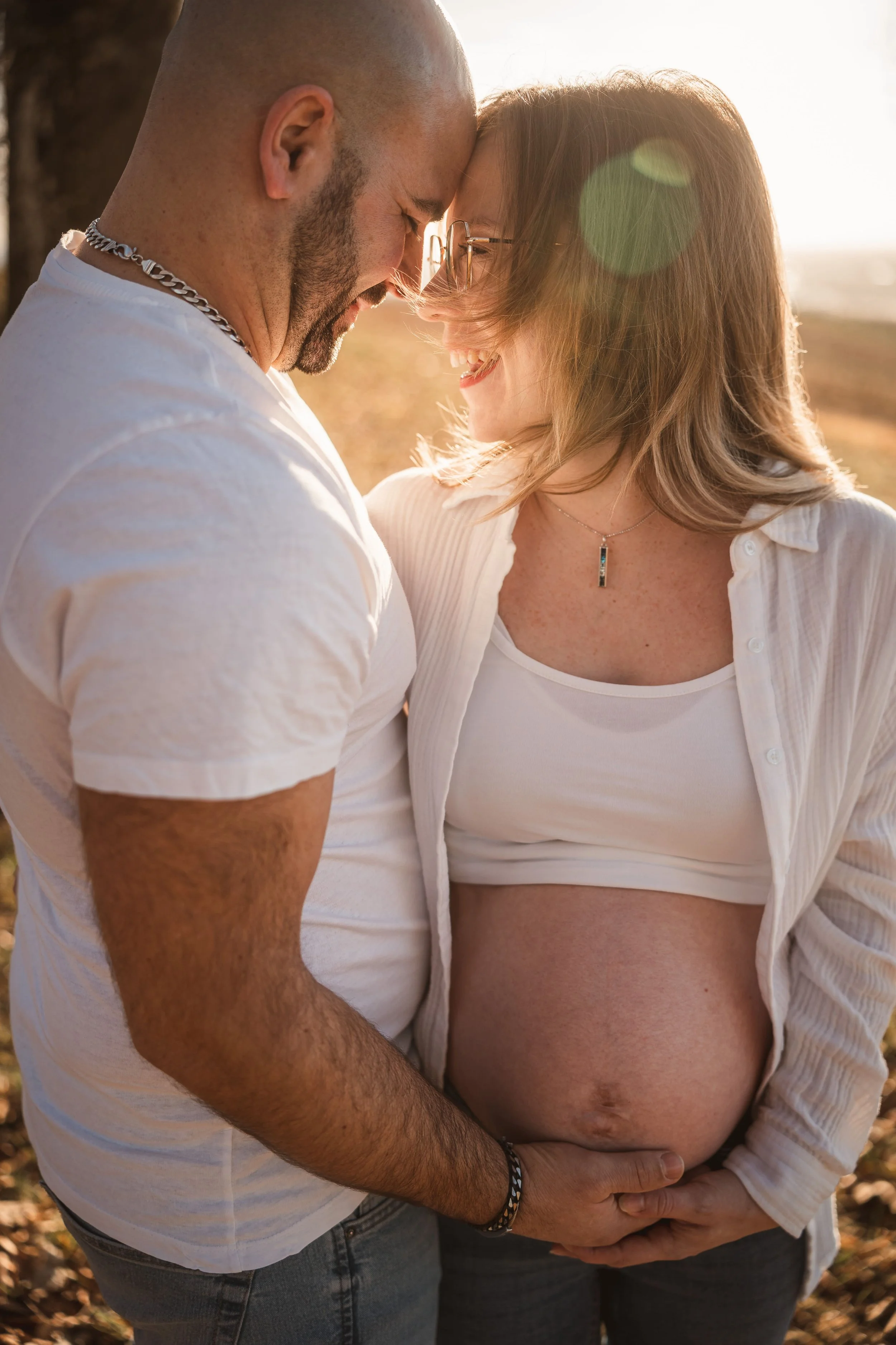 Ein Paar hält gemeinsam ihren Babybauch in der Natur bei Sonnenuntergang, lächelnd und nah beieinander.