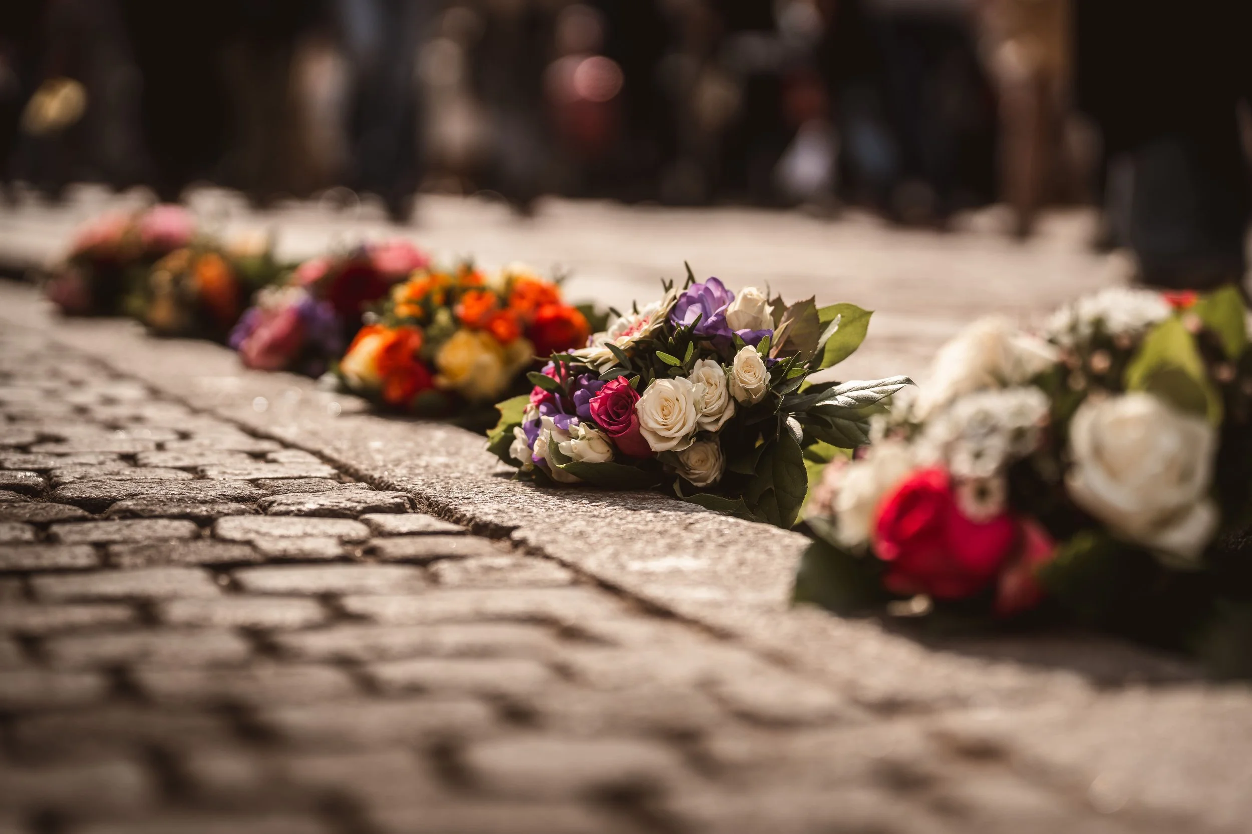 Blumen liegen auf einem Kopfsteinpflaster in einem Gedenken oder Kondolenzort, unscharfe Menschen im Hintergrund.