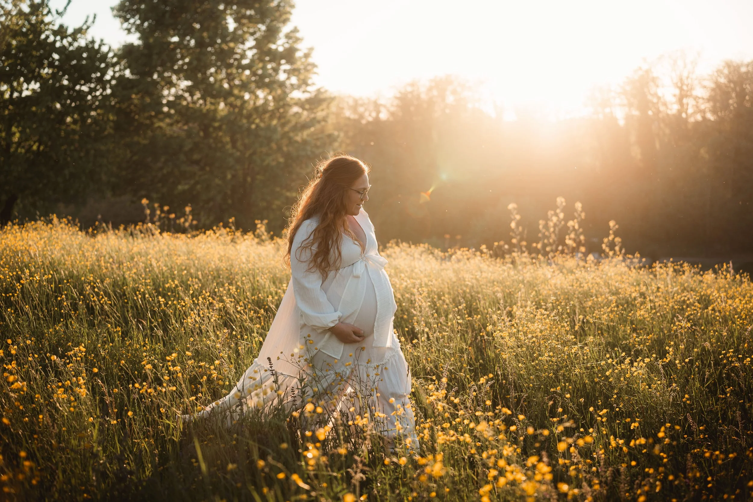 Eine schwangere Frau steht in einem blühenden Feld bei Sonnenuntergang, trägt ein weißes Kleid und lächelt sanft.