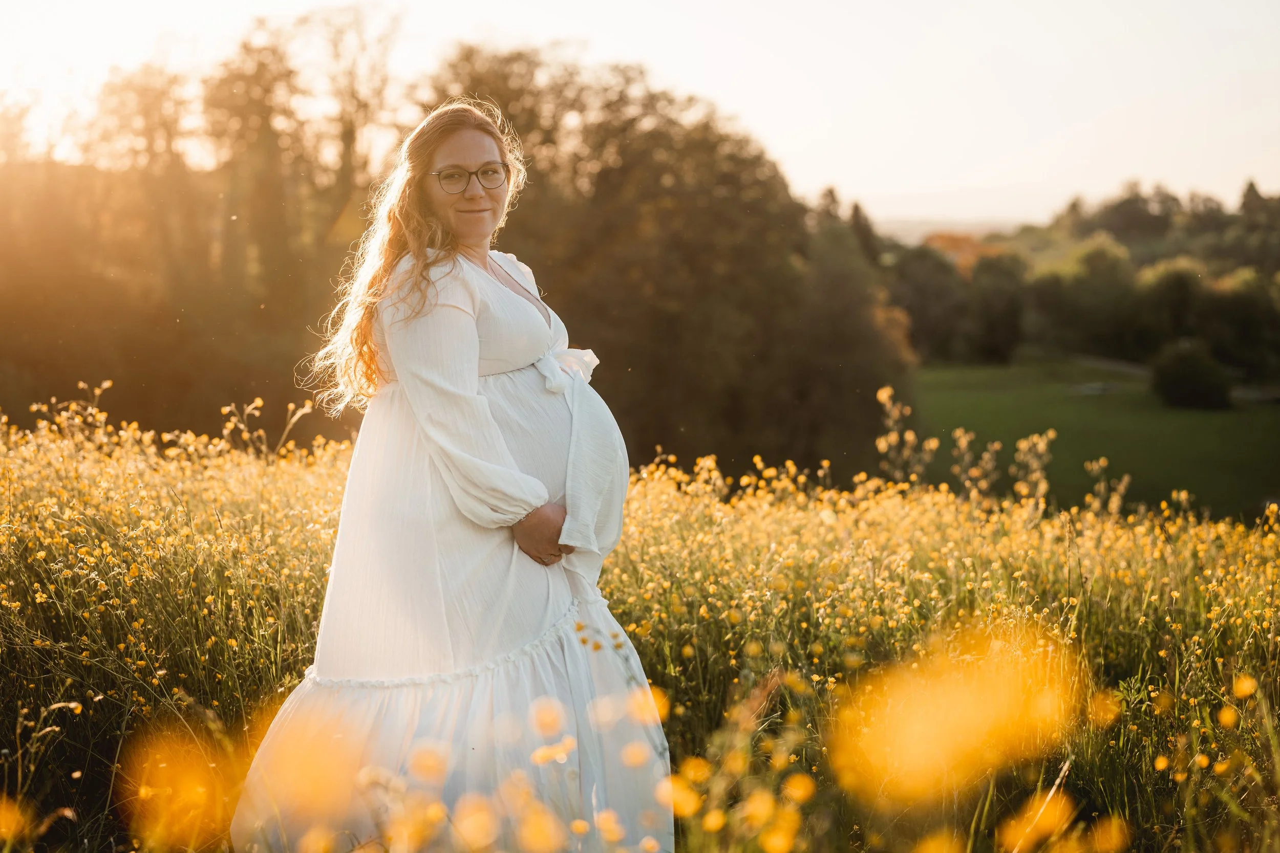 Schwangere Frau in weißem Kleid steht in einem Blumenfeld bei Sonnenuntergang, umgeben von gelben Blumen und Bäumen im Hintergrund.