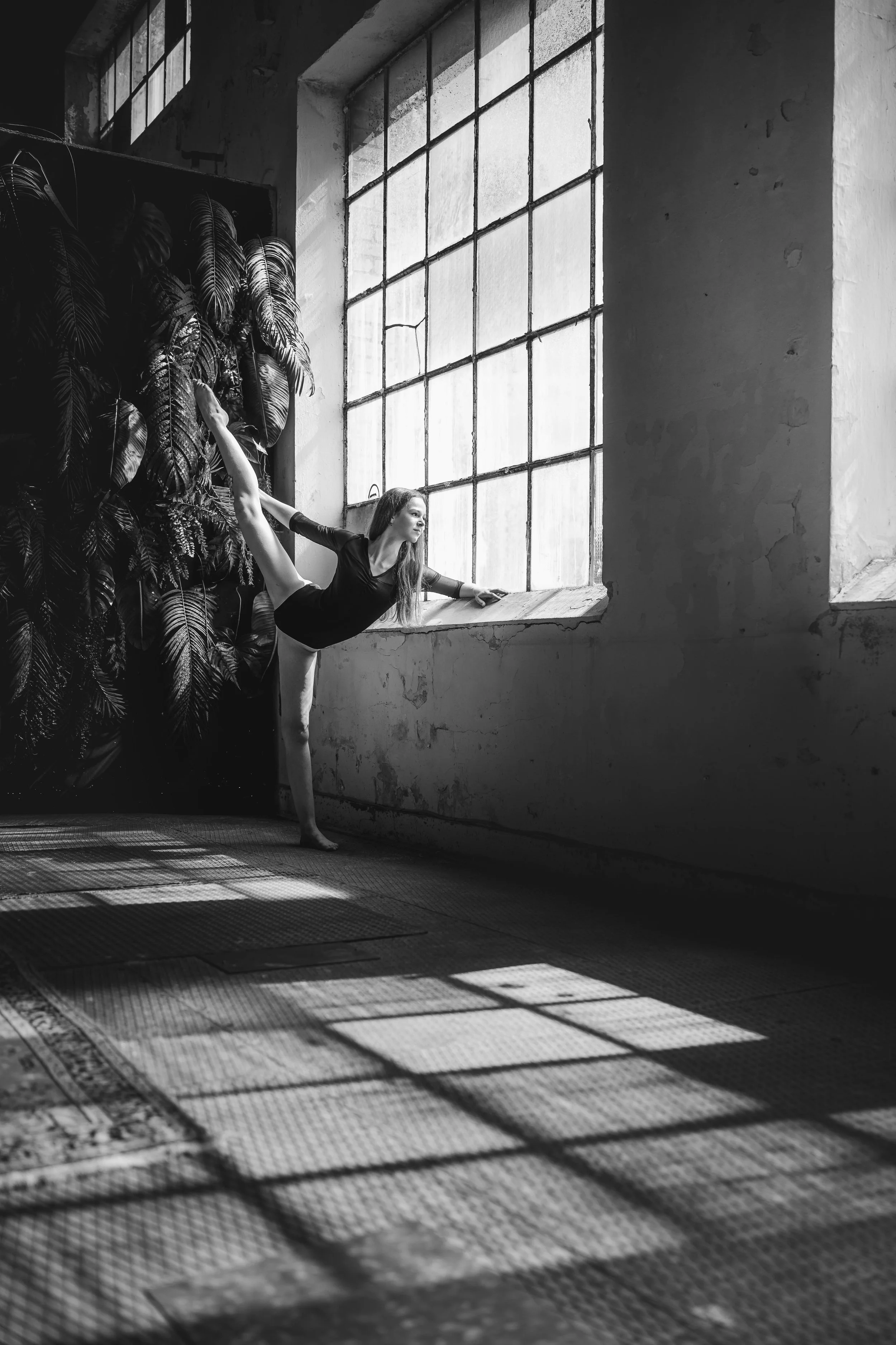 Schwarz-weiß-Foto eines jungen Mädchens, das Balletttanz übt, während es an einem Fenster in einem alten, industrialisierten Raum steht.