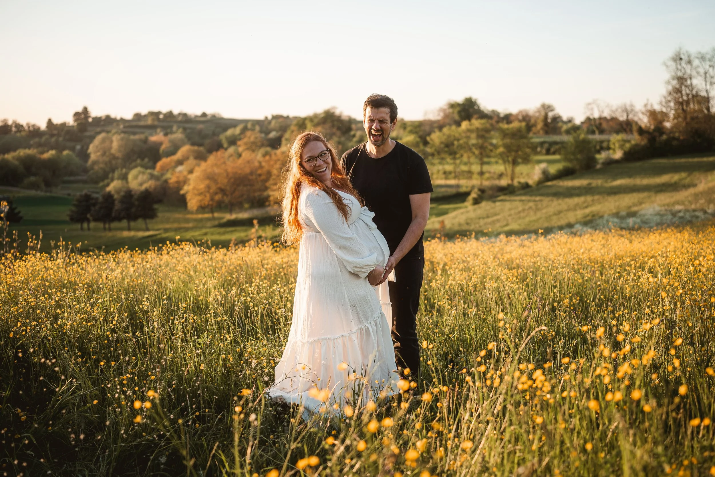 Ein schwangere Frau in einem weißen Kleid, lachend, zusammen mit einem lachenden Mann in einem schwarzen T-Shirt, stehen in einer Blumenwiese bei Sonnenuntergang, im Hintergrund Hügel und Bäume.