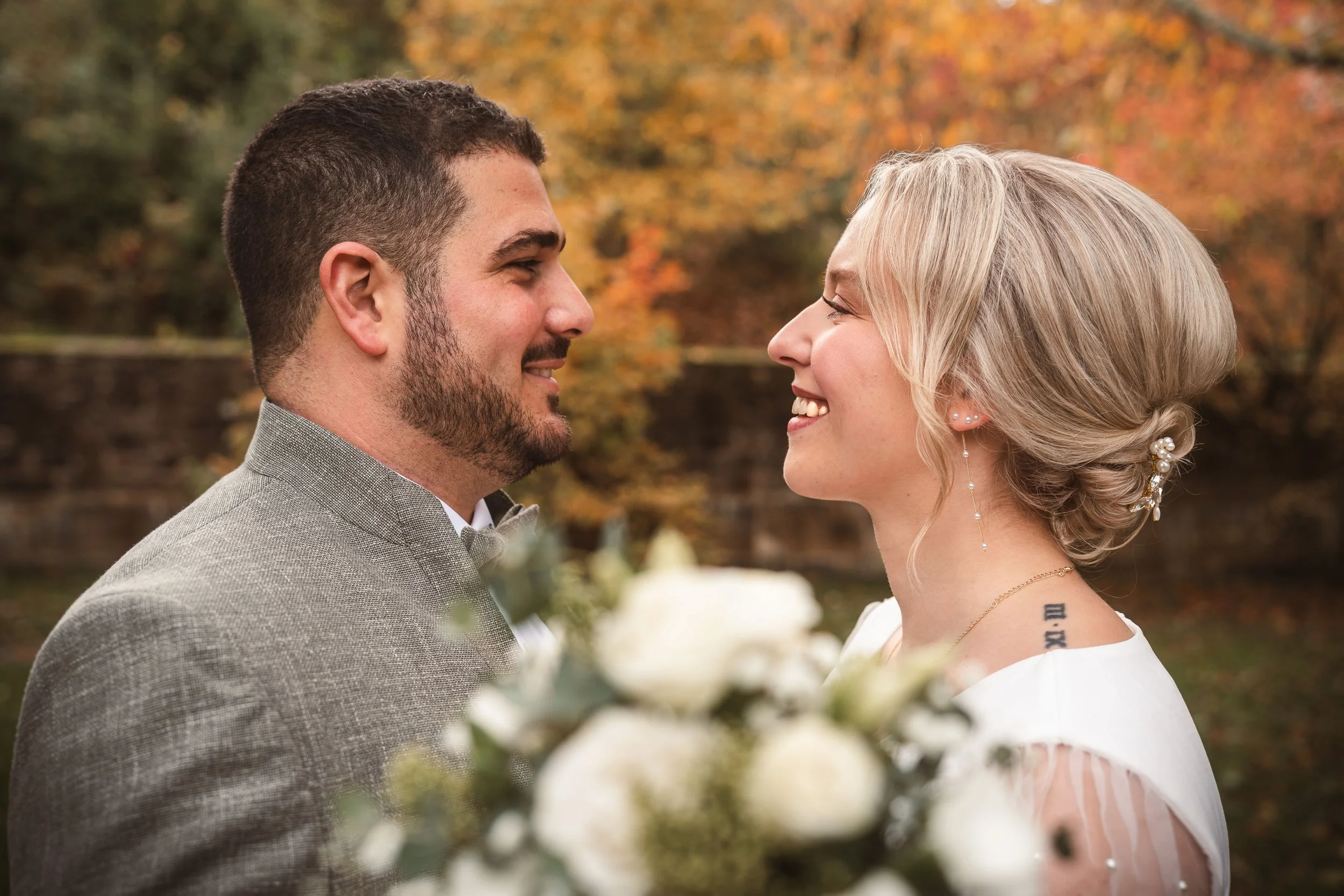 Ein verliebtes Paar, das sich gegenübersteht, im Freien bei Herbstlaub, lächelnd und Blick in die Augen. Die Frau trägt eine weiße Hochzeitsszene, der Hintergrund ist unscharf mit bunten Herbstblättern.