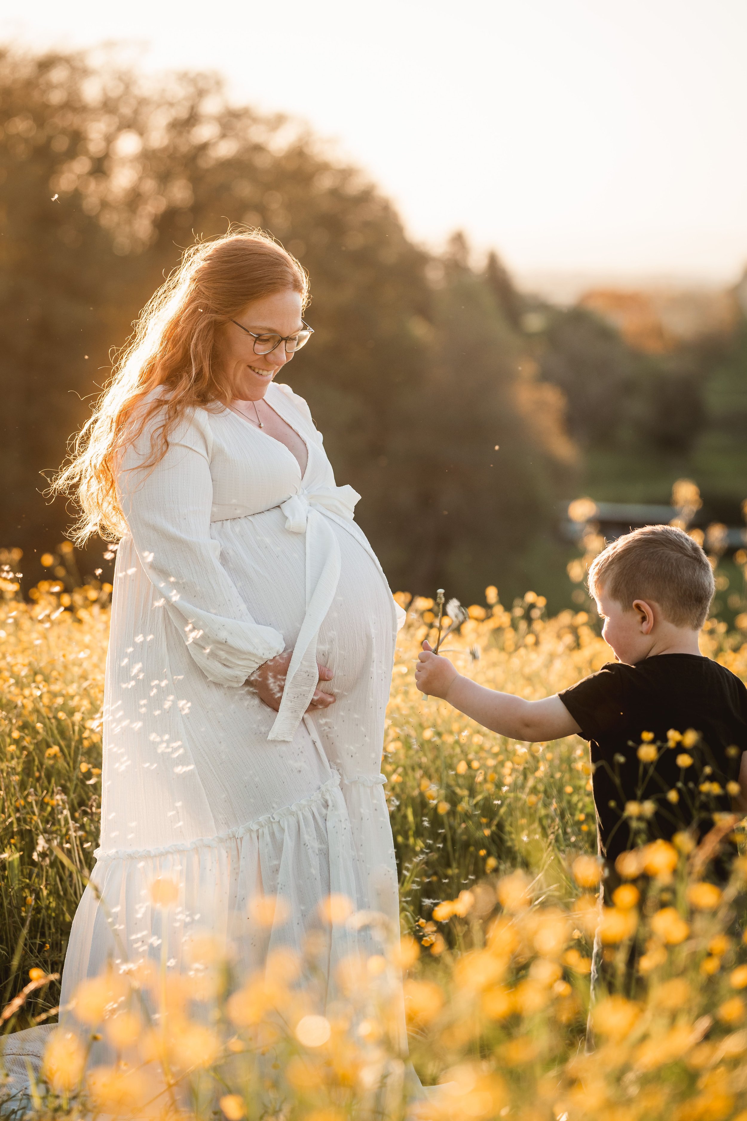 Schwangere Frau in weißem Kleid freut sich auf ein Treffen mit einem Jungen, während sie in einem Feld mit gelben Blumen im Sonnenuntergang steht.