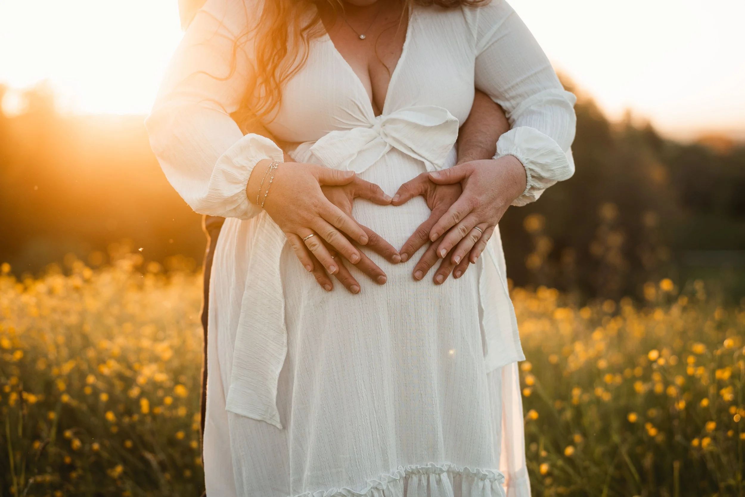 Eine schwangere Frau in weißem Kleid formt mit den Händen ein Herz auf ihrem Bauch, während sie im Sonnenuntergang in einem Feld voller gelber Blumen steht.