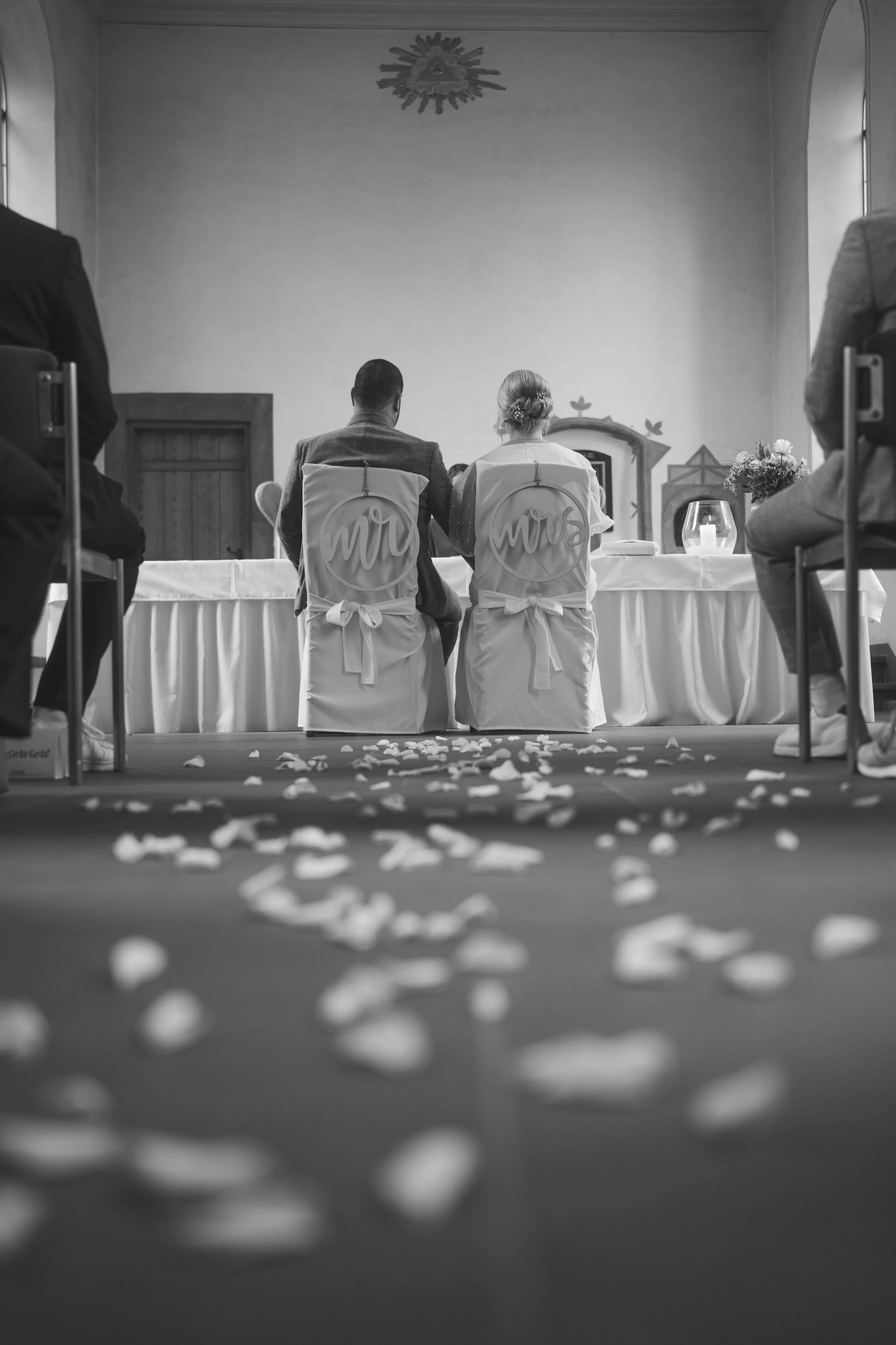 Brautpaar sitzt an einem Tisch bei einer Hochzeit, im Hintergrund Blumenschmuck und Wanddekoration, auf dem Boden liegen Blütenblätter.