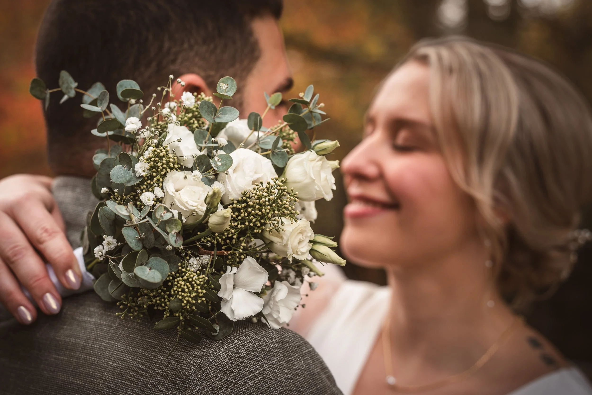 Ein Paar umarmt sich bei einer Hochzeit, die Braut hält einen Blumenstrauß mit weißen Blumen und grünen Blättern. Das Foto ist nah aufgenommen und zeigt nur Teile der Personen.