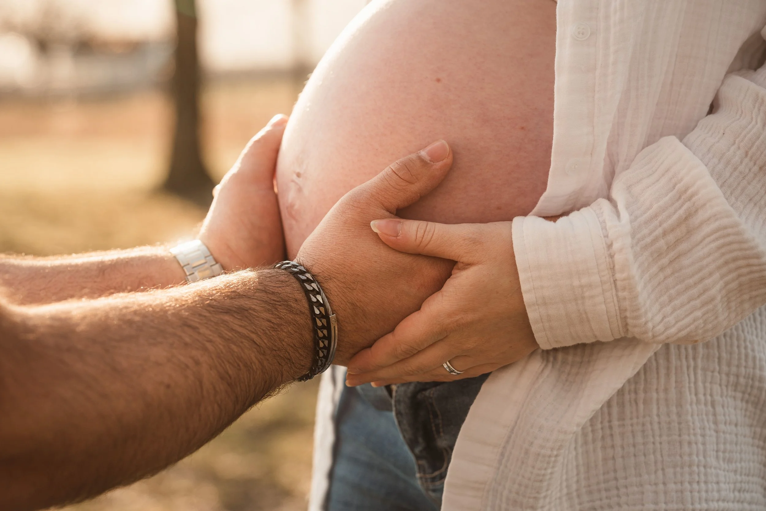 Nahaufnahme einer Hand, die einen Bauch berührt, im Hintergrund eine weitere Person, die ihren Bauch berührt, bei Sonnenlicht