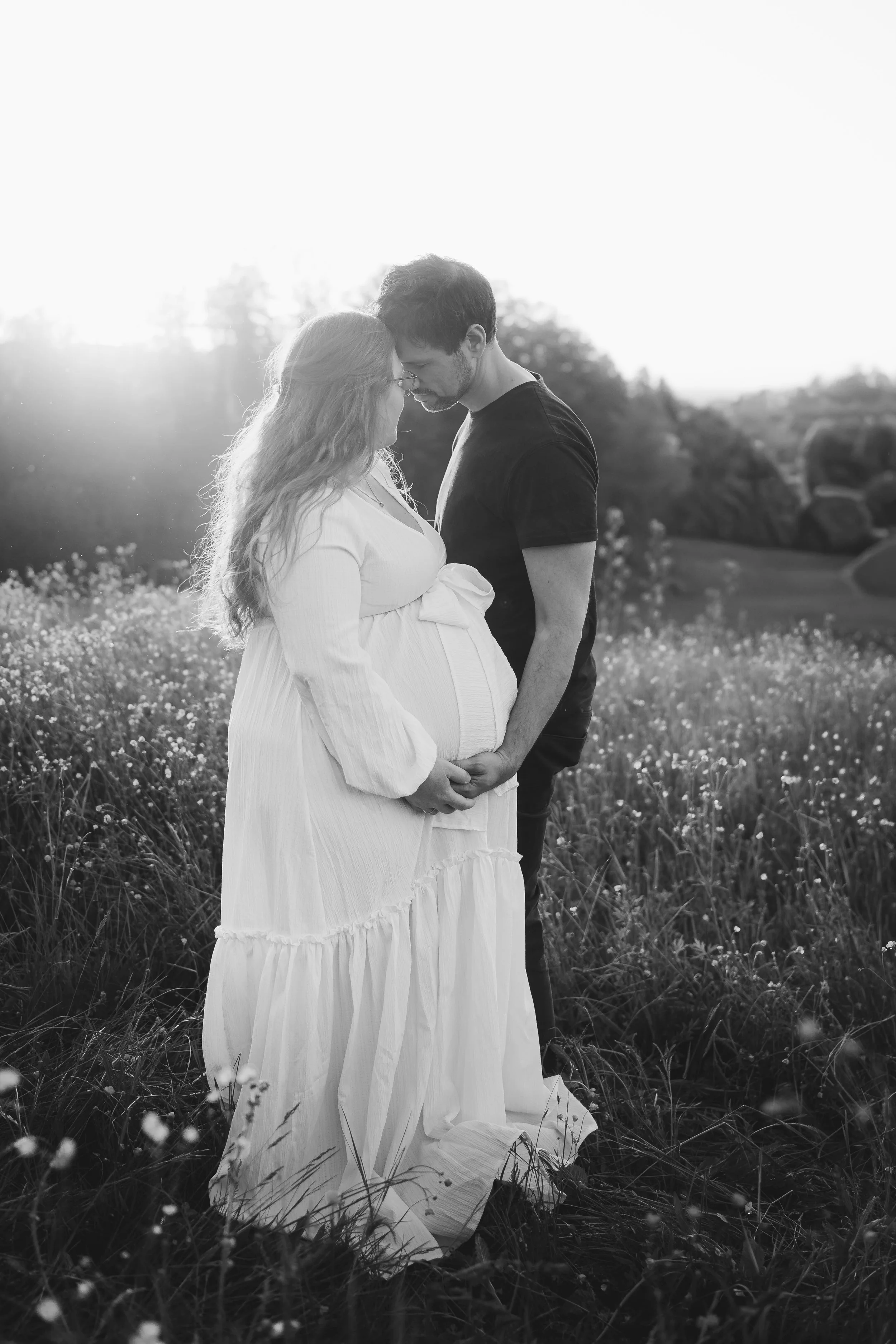 Ein schwaz-weißes Bild eines Paares in einem Blumenfeld, das sich liebevoll aneinander lehnt. Die Frau ist schwangere und trägt ein langes weißes Kleid, der Mann trägt ein dunkles T-Shirt.