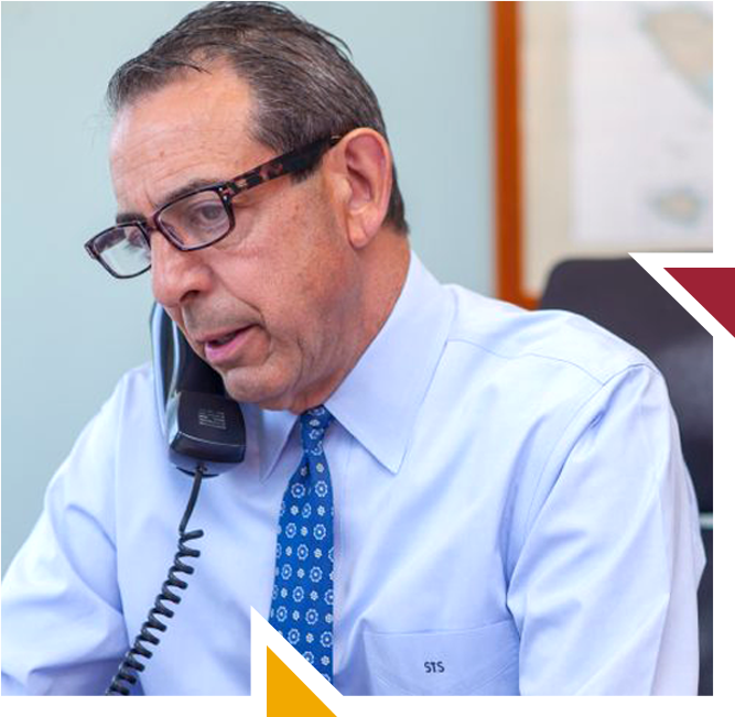 A middle-aged man with glasses, wearing a light blue dress shirt and patterned tie, talking on a landline telephone in an office setting.