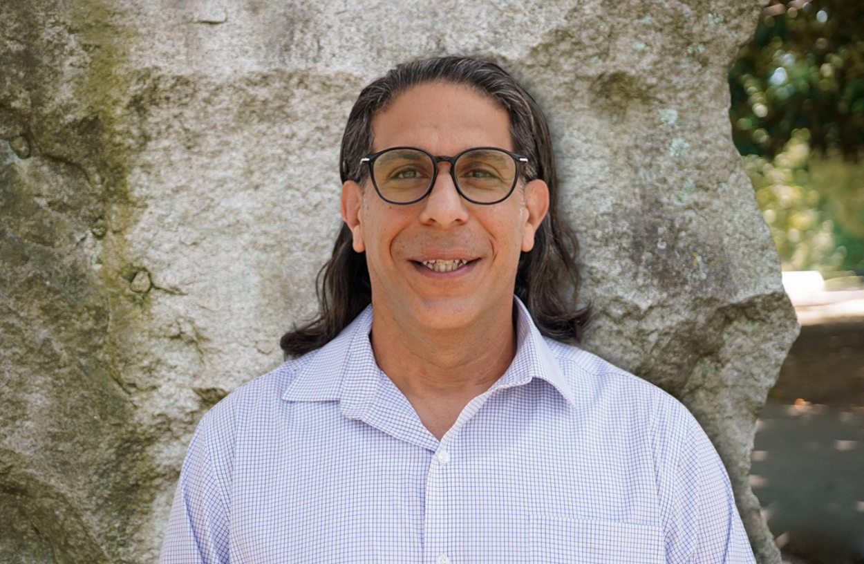 Aurang of Grovestone Countertops. A man with long dark hair, glasses, and a white checkered shirt standing outdoors in front of a large rock.