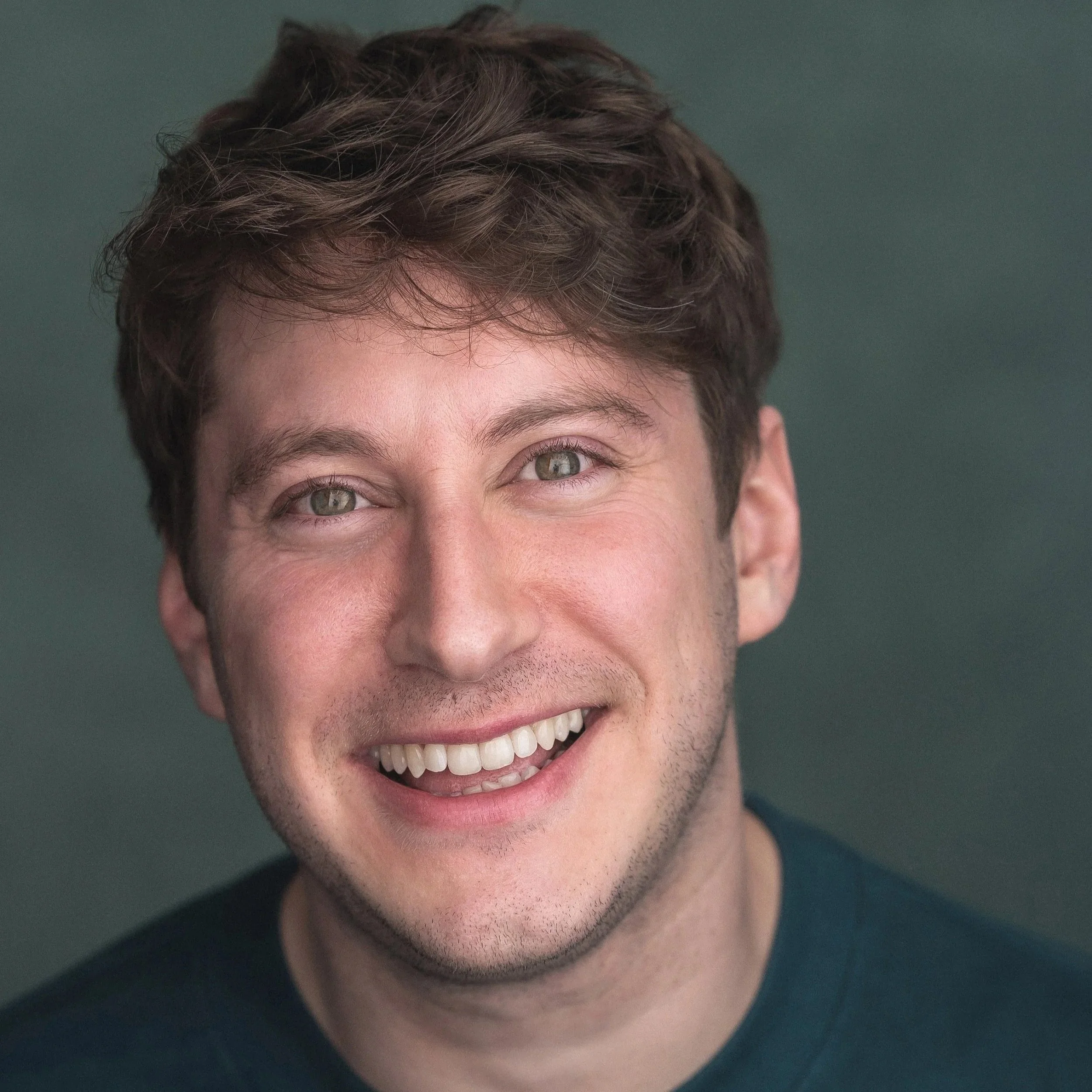 Close-up of a young man with short, wavy brown hair, light green eyes, and a big smile showing his teeth, wearing a dark shirt against a plain gray background.