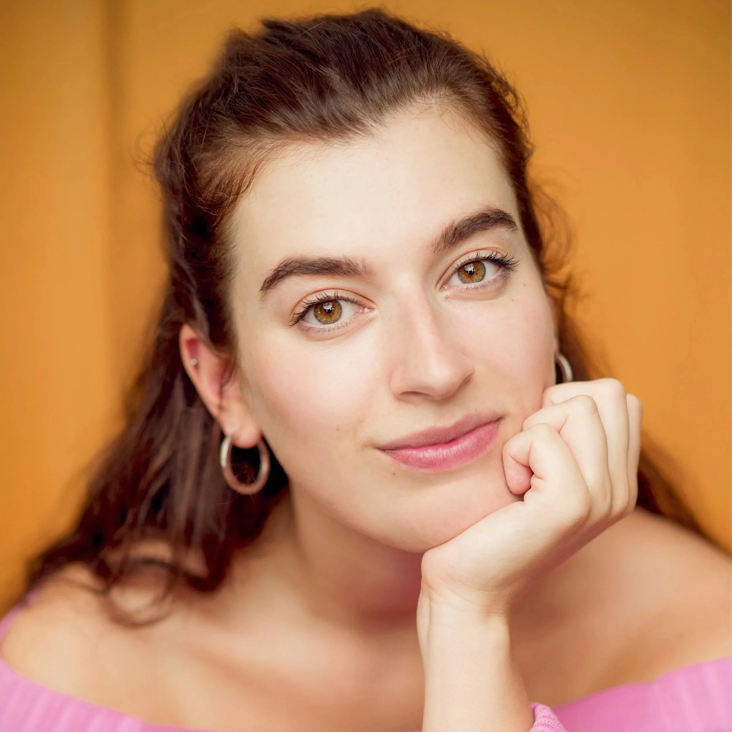 Close-up of a young woman with brown hair and green eyes resting her chin on her hand against an orange background.