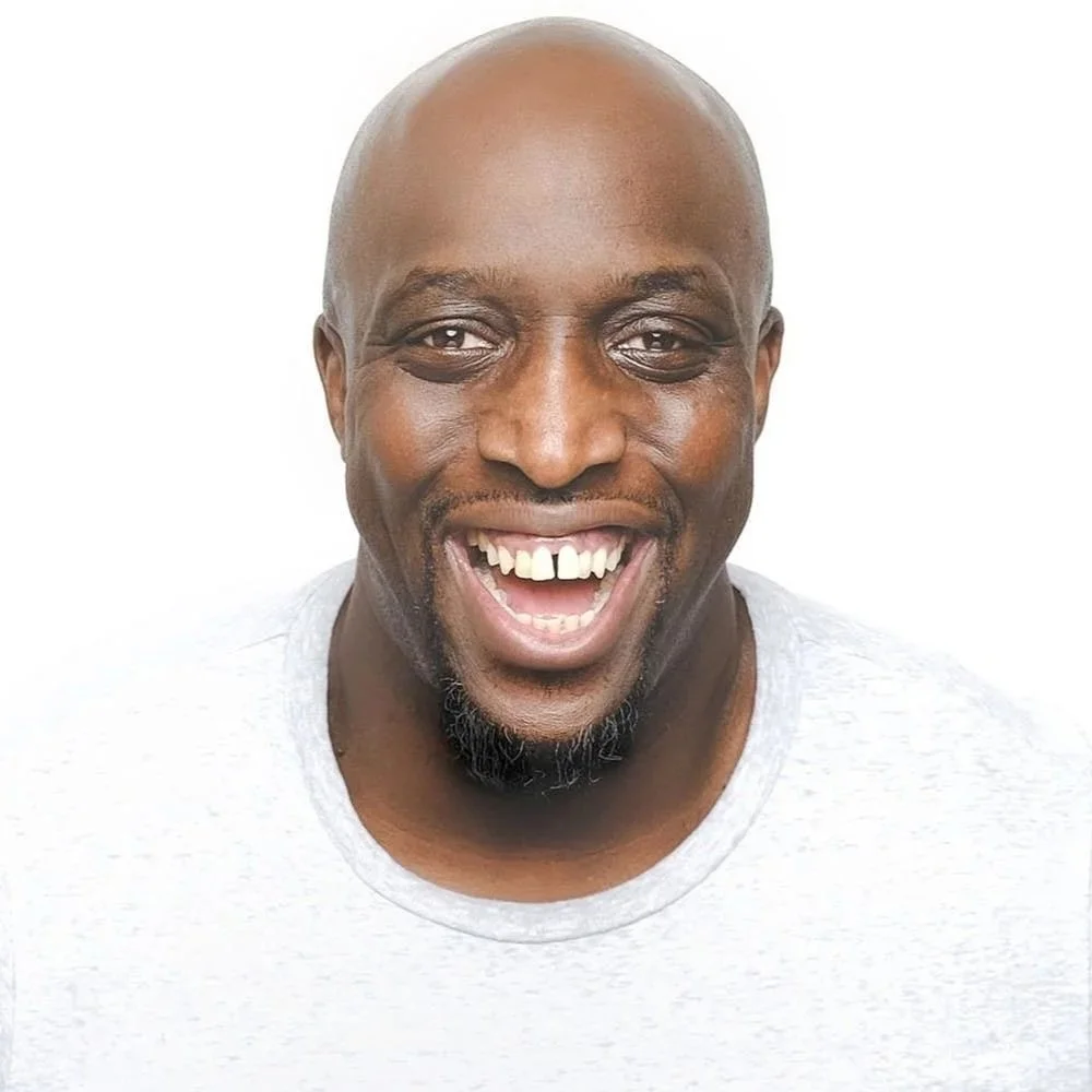 A smiling African American man with a bald head and a goatee, wearing a light gray shirt, against a white background.