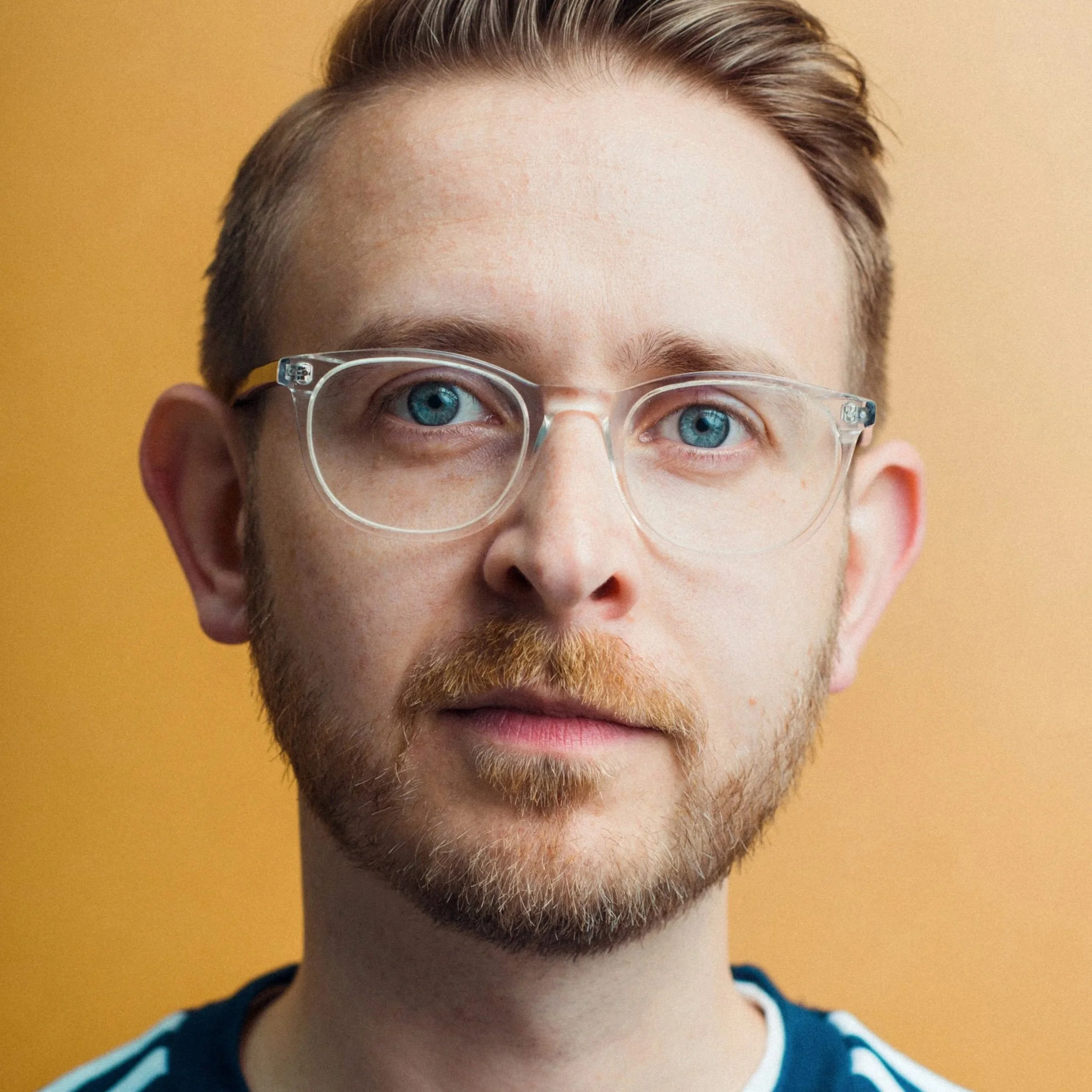 Portrait of a young man with light skin, glasses, and a short beard, against a tan background.