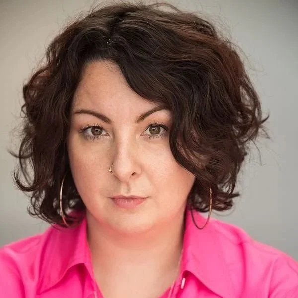 Close-up portrait of a woman with short, curly dark hair wearing a bright pink shirt and large hoop earrings.