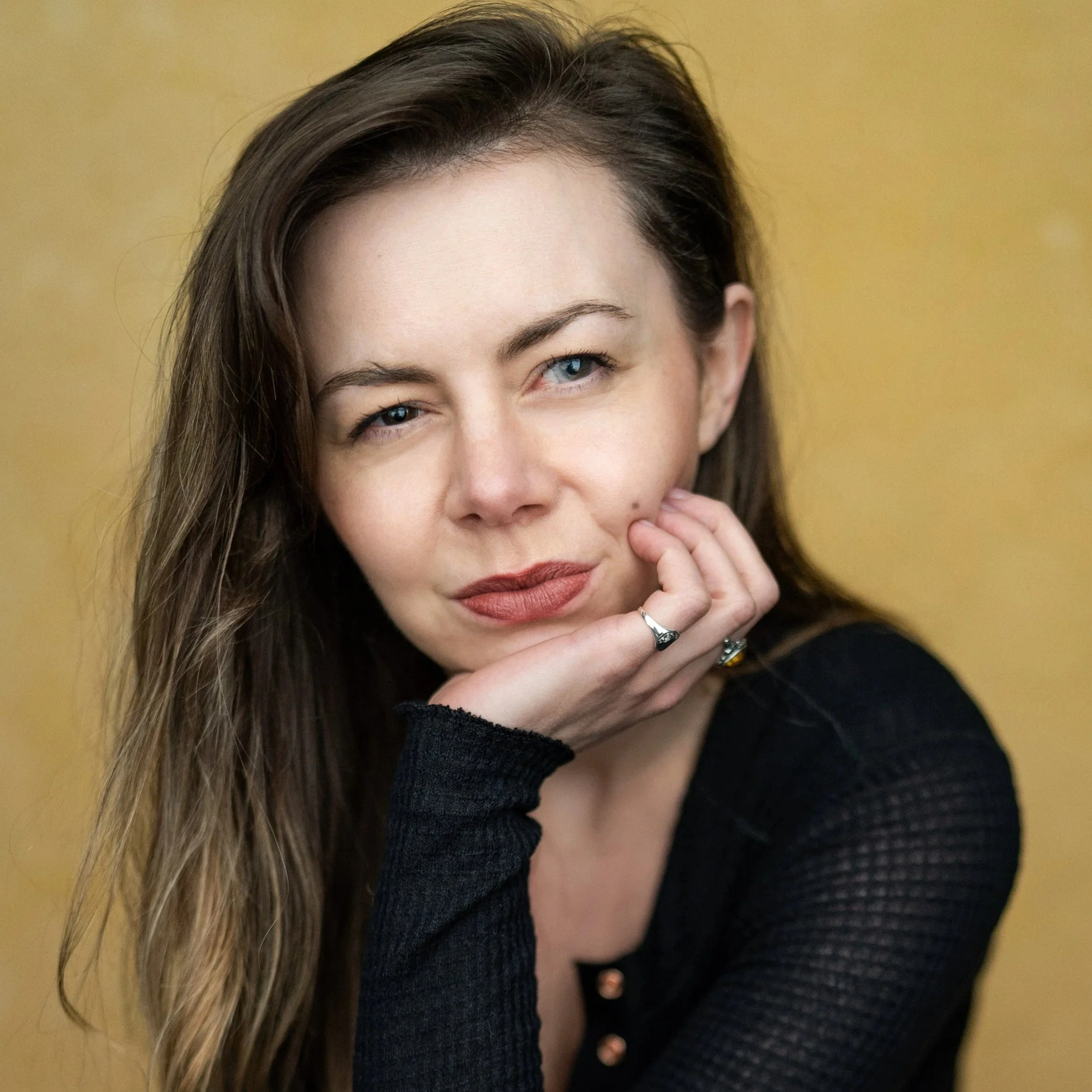 Portrait of a woman with long brown hair and light skin, resting her chin on her hand, wearing a black top, against a yellow background.
