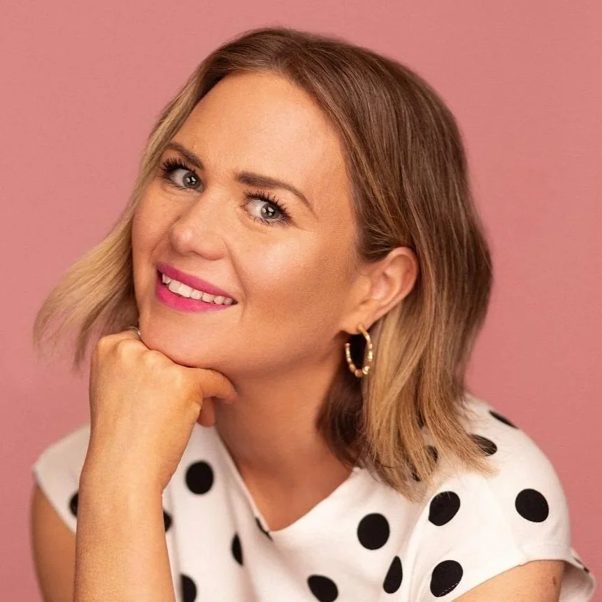 A woman with shoulder-length blonde hair, wearing a white shirt with black polka dots and gold hoop earrings, smiling and resting her chin on her hand against a pink background.