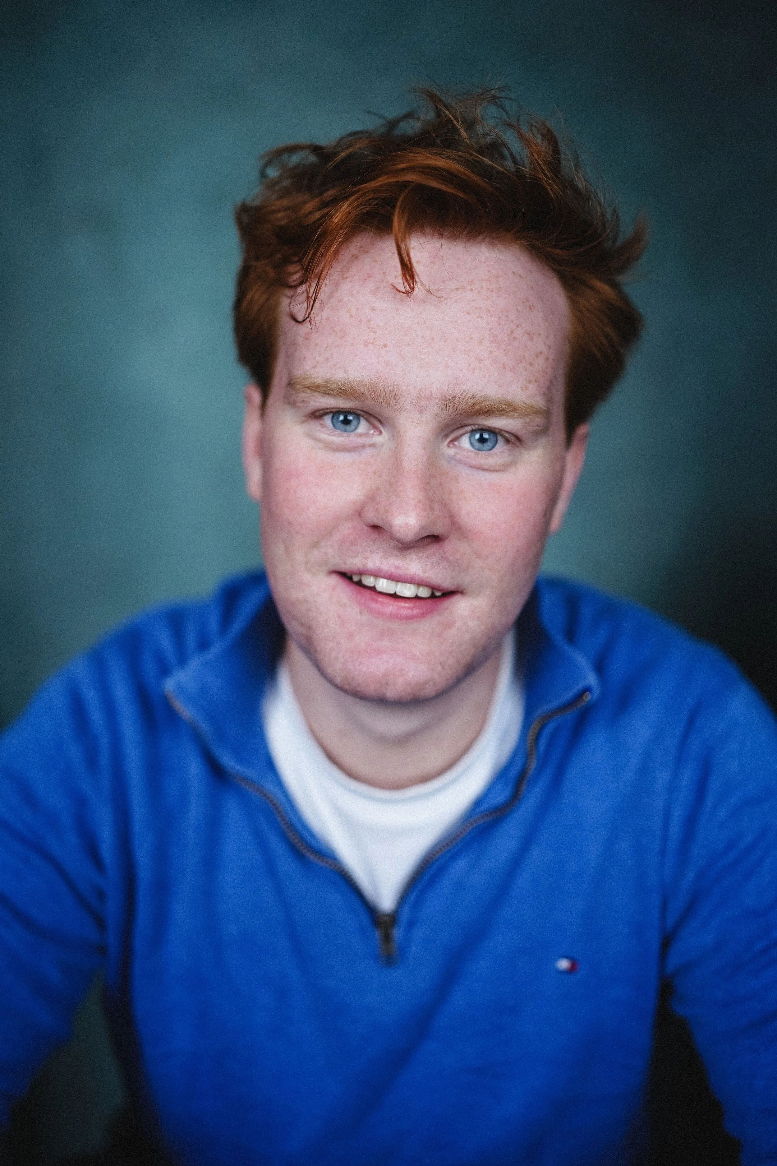 Headshot of a young man with red hair and blue eyes, smiling, wearing a blue zip-up jacket over a white t-shirt.