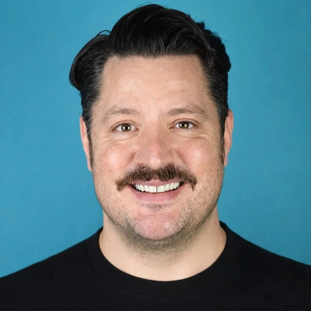 Close-up portrait of a smiling man with brown eyes, dark hair, and light skin, wearing a blue shirt.