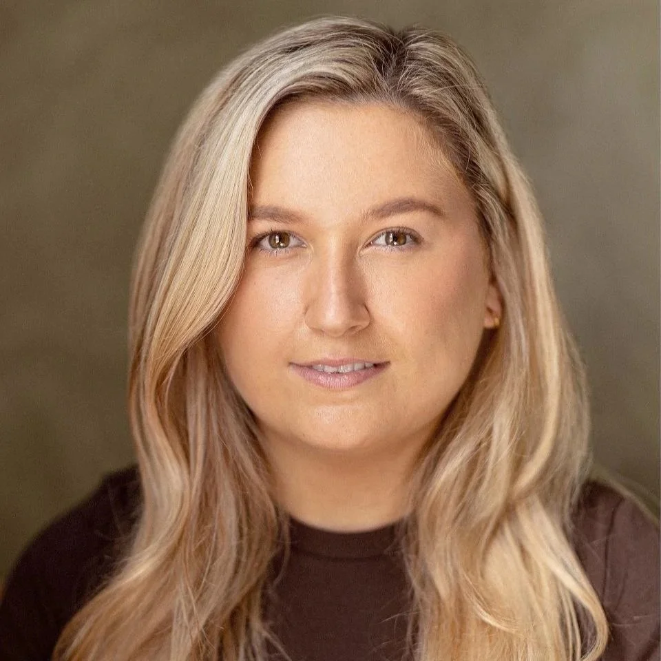 Close-up portrait of a young woman with long blonde hair, light makeup, and a neutral expression, wearing a dark top.