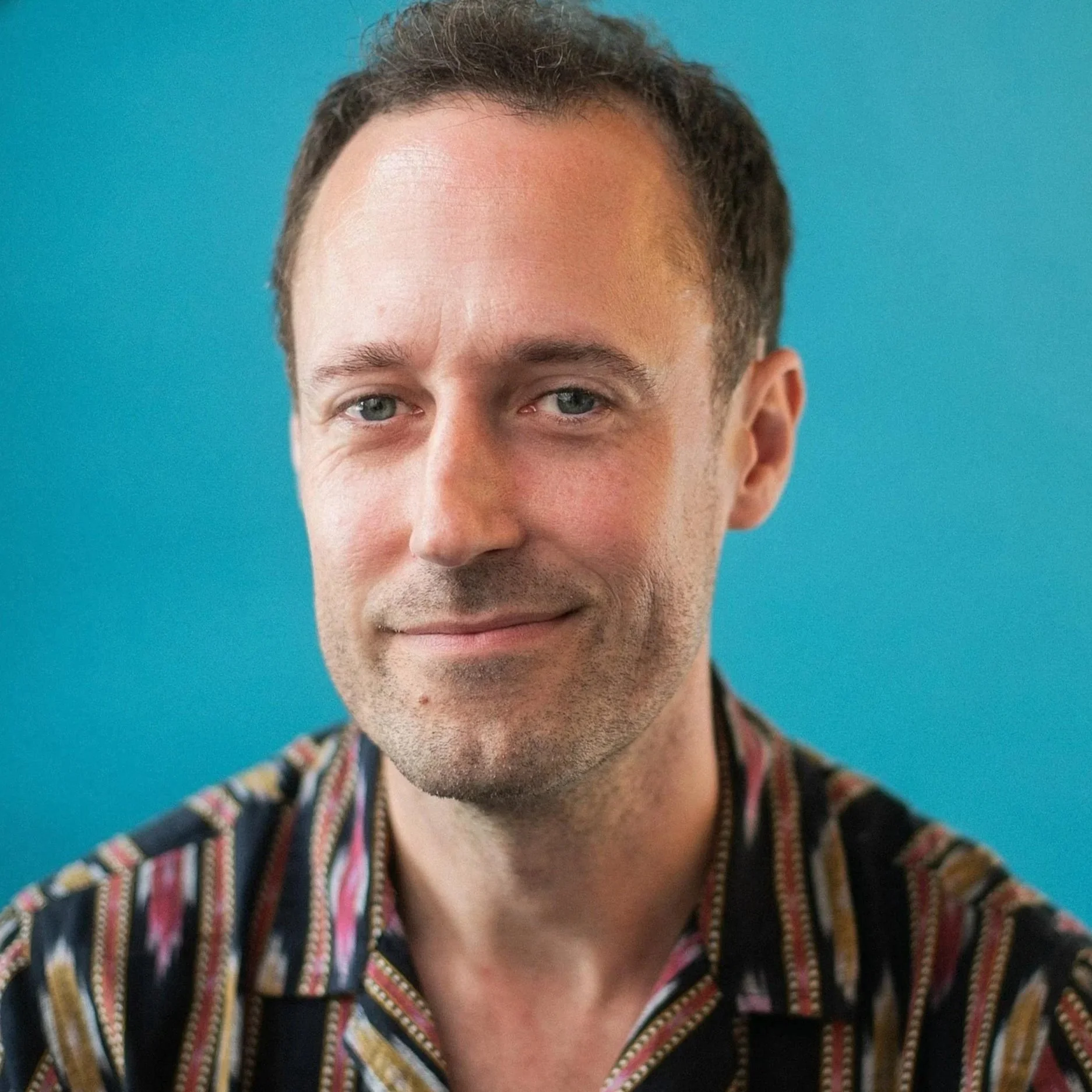 A man with short brown hair, blue eyes, and a light beard wearing a dark blue shirt against a dark background.