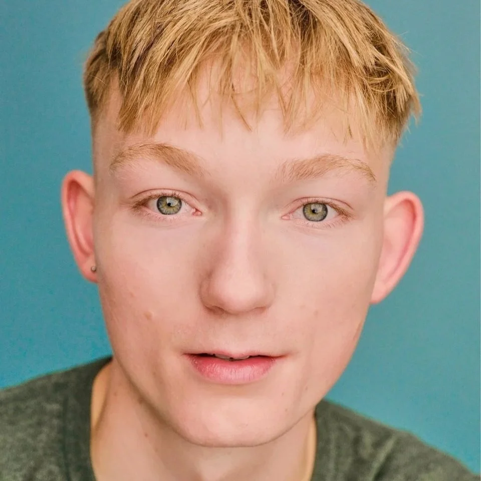 Close-up of a young person with short blond hair, light skin, and green eyes against a teal background.