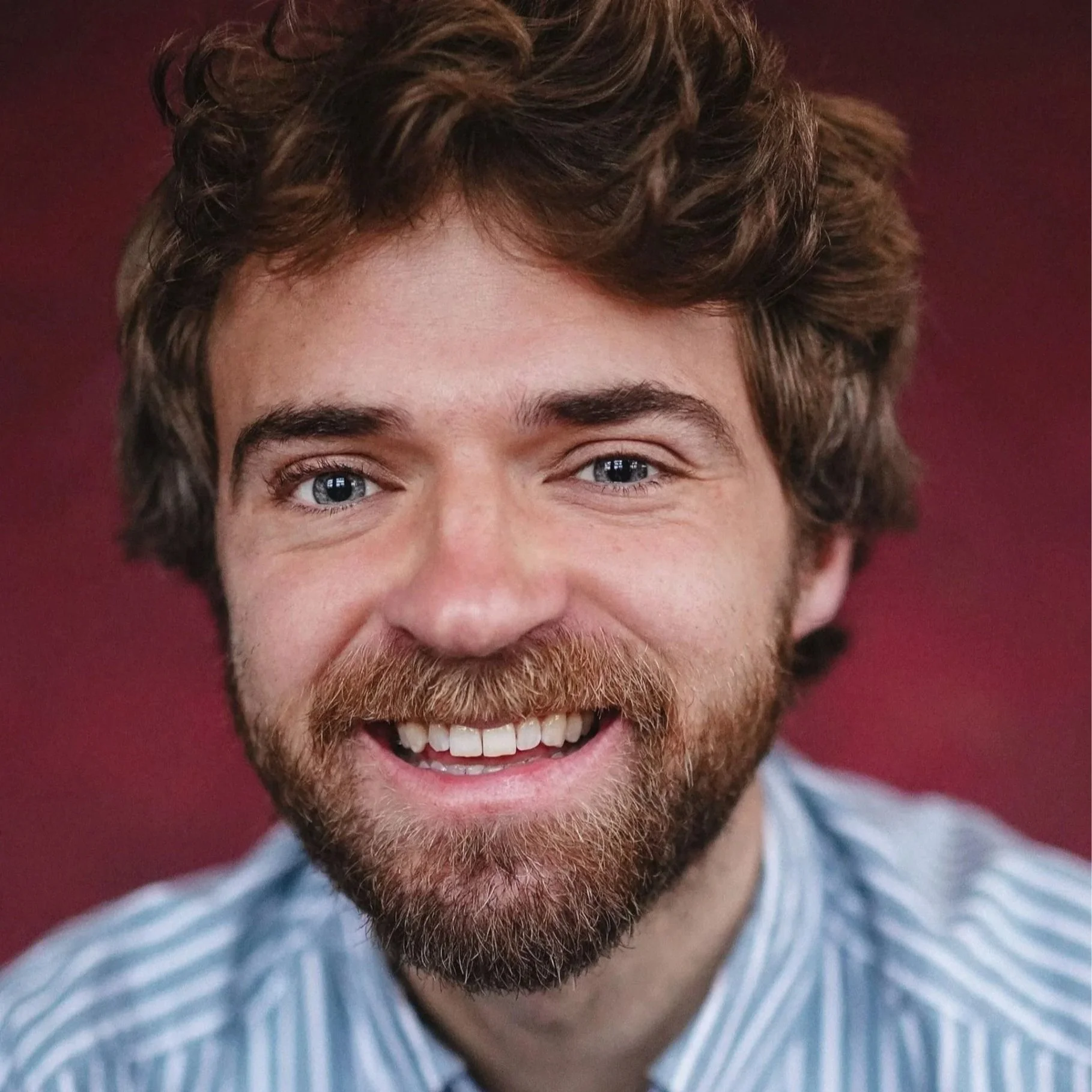 Close-up of a smiling man with blue eyes, brown curly hair, and a beard, wearing a light blue and white striped shirt, against a maroon background.