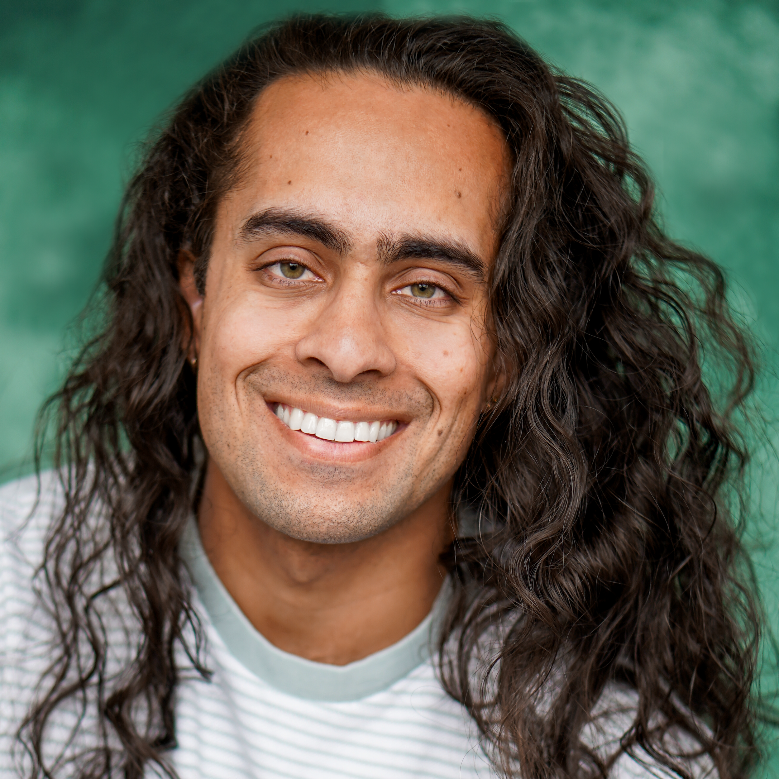 Close-up of a smiling man with long, curly dark hair and light-colored eyes, wearing a white and light green striped shirt, in front of a green background.