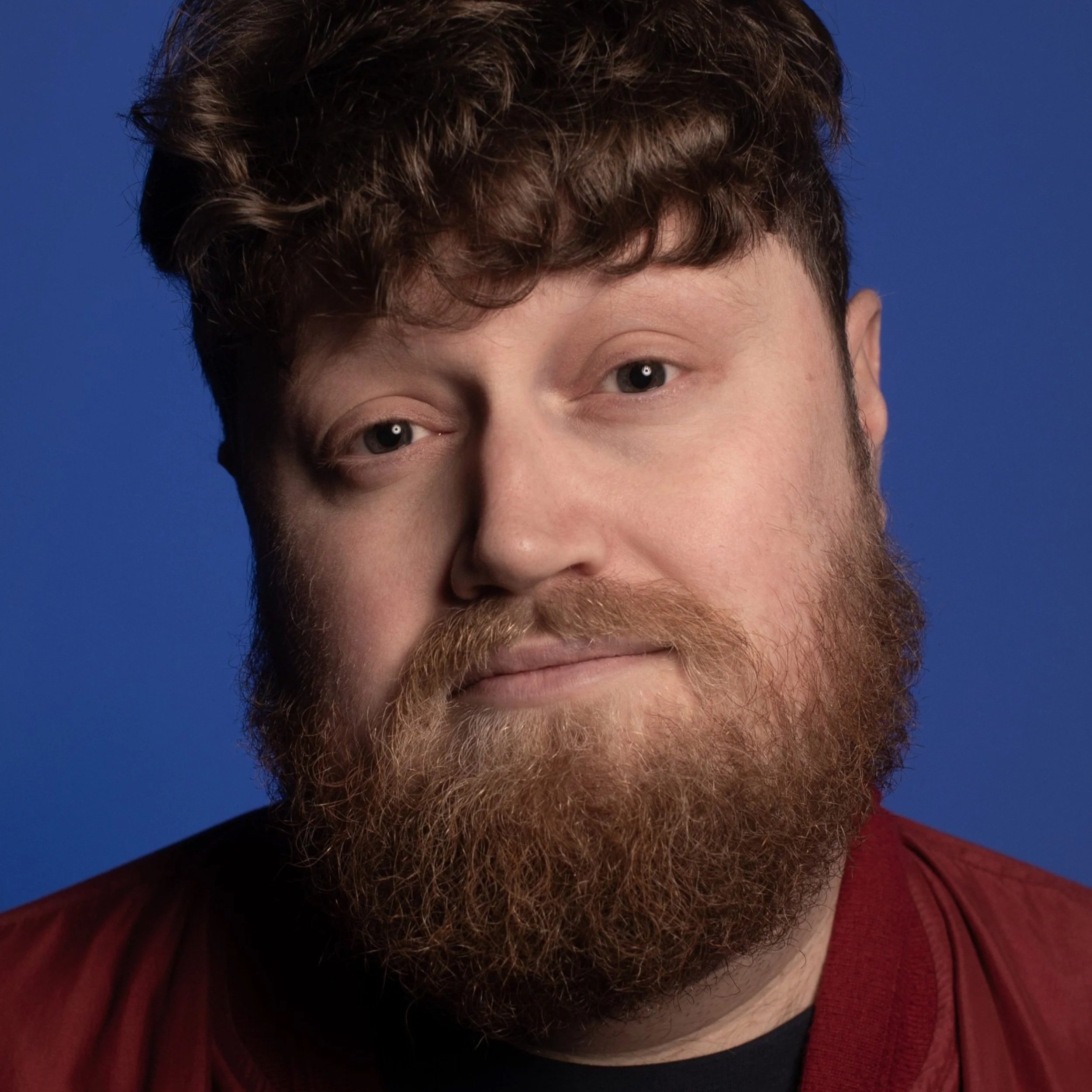 Close-up portrait of a man with curly brown hair, a full beard, and blue eyes, wearing a maroon jacket against a solid blue background.
