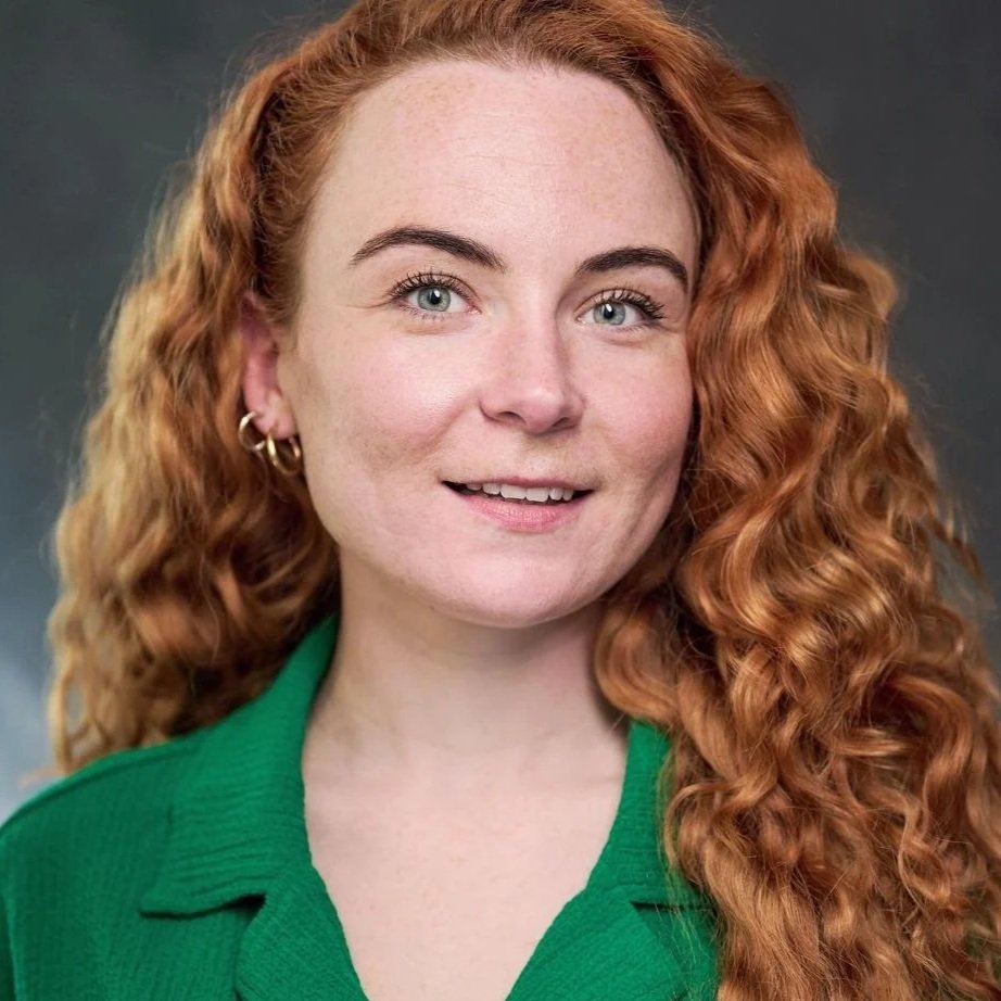 Close-up portrait of a smiling woman with curly red hair, blue eyes, and hoop earrings, wearing a green top, against a neutral gray background.