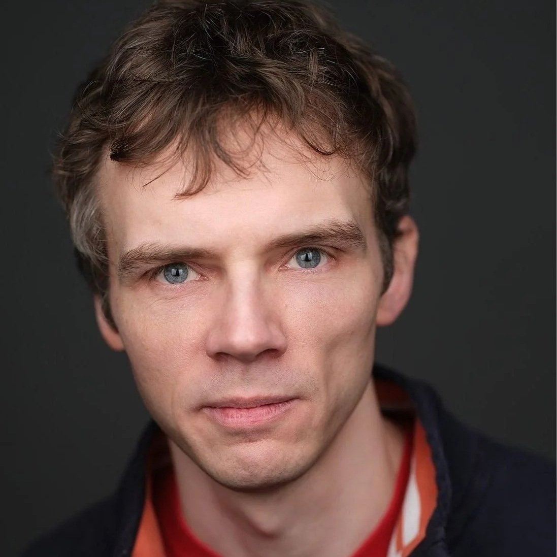 Close-up portrait of a young man with blue eyes, light brown hair, and fair skin, wearing a navy hoodie over a gray shirt, smiling slightly at the camera.